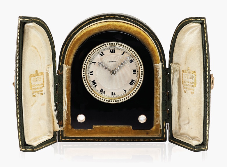 An Art Deco onyx, moonstone and enamel desk clock, 1920s, by Cartier. The circular cream engine-turned dial with painted black Roman numerals, outer minute railway and rose-cut diamond-set arrow-shaped hands, within a white enamel bezel, to the onyx stand enhanced by two cabochon moonstones, mechanical movement, 1920s, 7.5 cm, in green leather original Cartier case with stand. Estimate