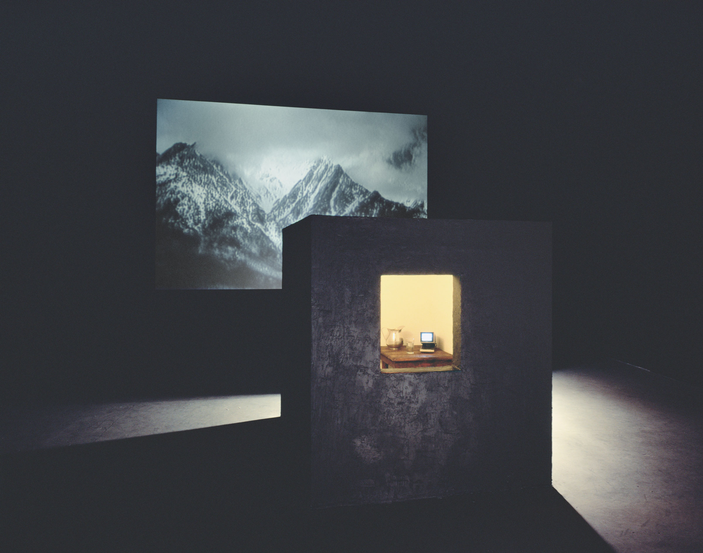 Bill Viola, Room for St. John of the Cross, 1983. Video and sound installation in a dark room, with a black cubicle with window, peat moss, wooden table, glass and metal pitcher with water, colour video, and one-channel mono sound; black-and-white video projection; amplified stereo sound; room: 14 x 24 x 30 ft (4.3 x 7.3 x 9.1 m); projected image: 8 ft 7 in x 12 ft 8 in (2.6 x 3.7 m); continuously running. Photo: Kira Perov