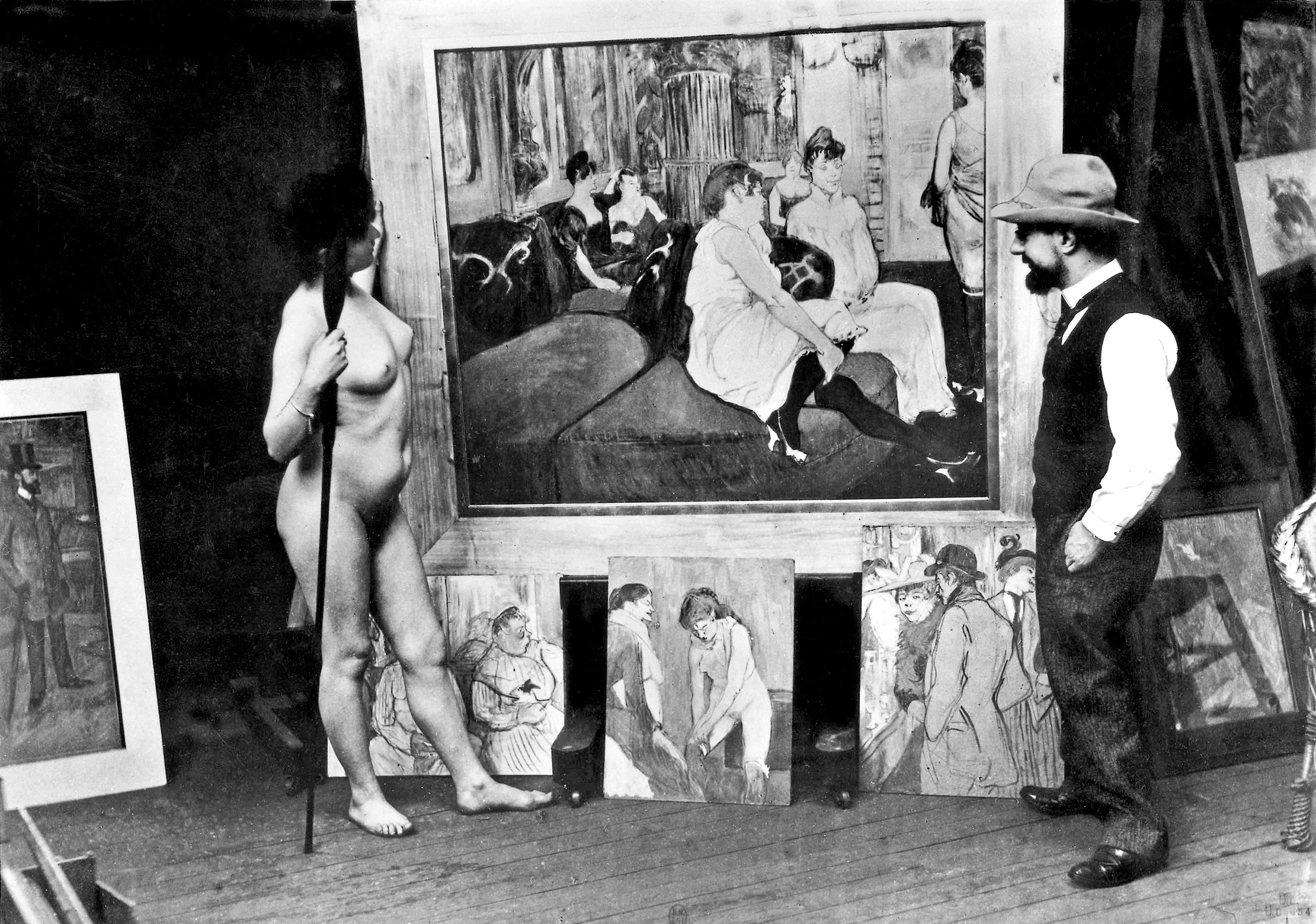 A photograph of Toulouse-Lautrec and a model with several of his works, including the 1894 paintings Au salon de la rue des Moulins and Femme tirant son bas