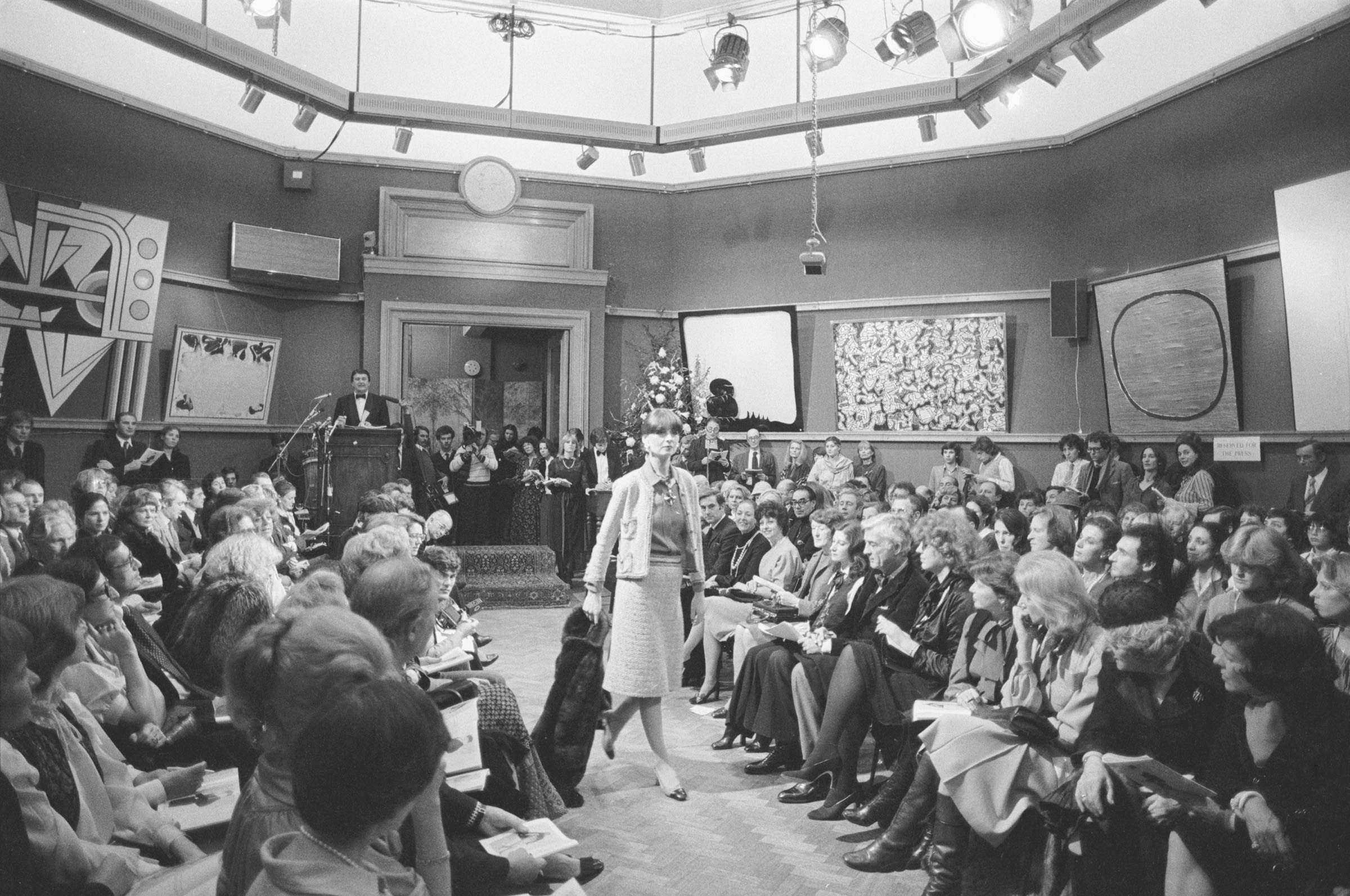 Modelling a tweed suit and a mink coat at the Chanel auction on 2 December 1978 at Christie's in London. On the walls are works from the 6 December Contemporary Art sale, from left: Roy Lichtenstein, Modern Painting No. 1, 1966; Sam Francis, Composition in Blue and White, 1960; Lucio Fontana, Concetto Spaziale, 1965; Jean Dubuffet, Banc de Prosperite, 1963; and Lucio Fontana, Laguna di Venezia, 1956