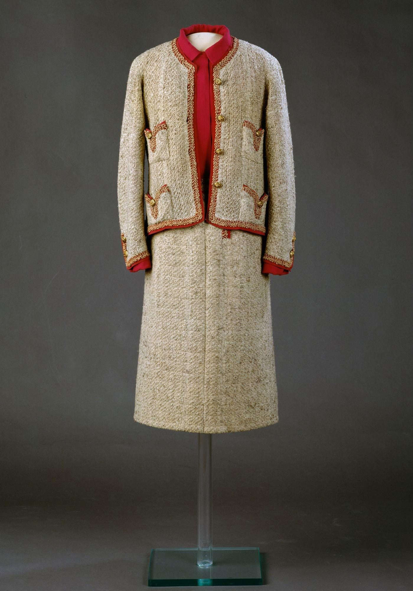 A suit of beige tweed bound with braid and bright pink silk, the pockets dipping in the middle with gilt lions' head buttons, weighted with chains, with a blouse of cyclamen pink silk crepe. Chanel designed this suit for herself and wore it often, particularly at the time of her last three collections