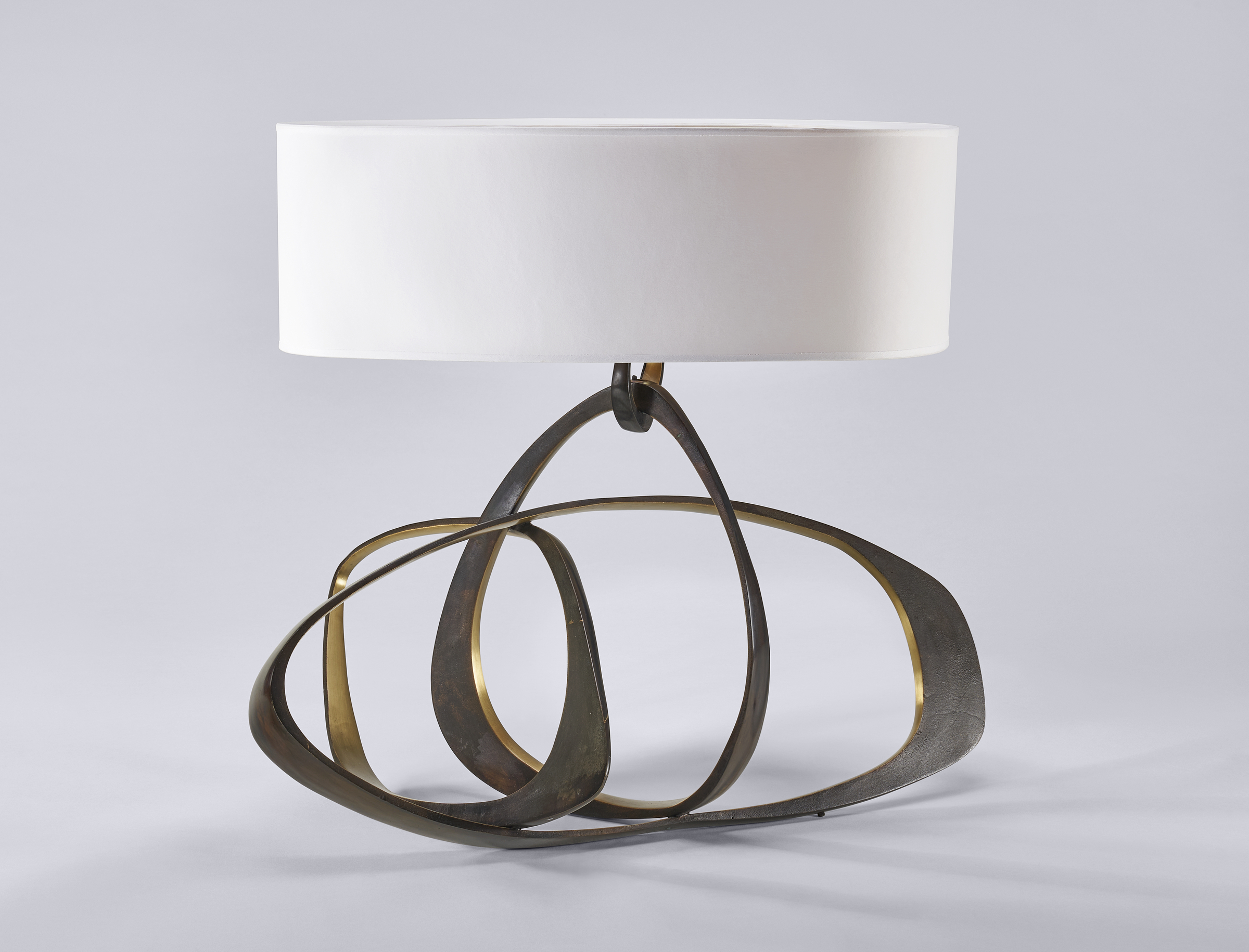 A modern table lamp featuring a circular, intertwined metallic base and a plain white shade.