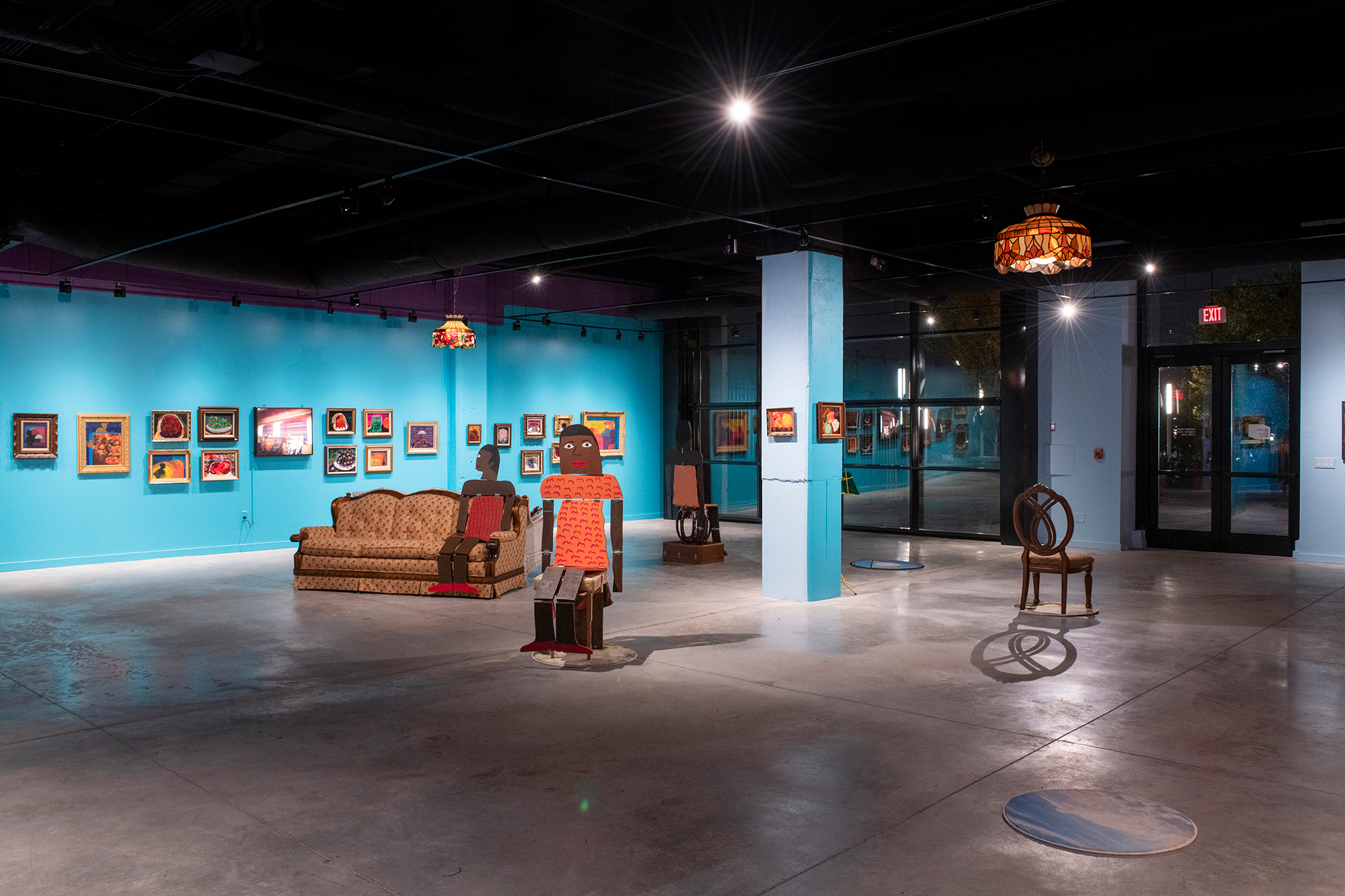 Art gallery interior with bright turquoise walls, colorful framed art, eclectic furniture, and whimsical wooden figures.