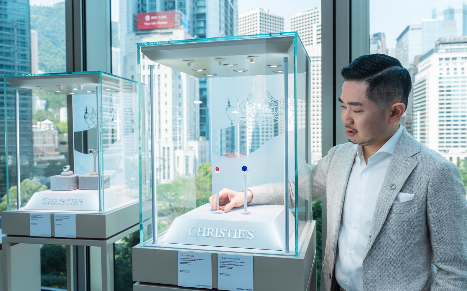 A person in a gray suit stands beside glass display cases in a room overlooking a cityscape, showcasing elegant items with Christie’s labels.