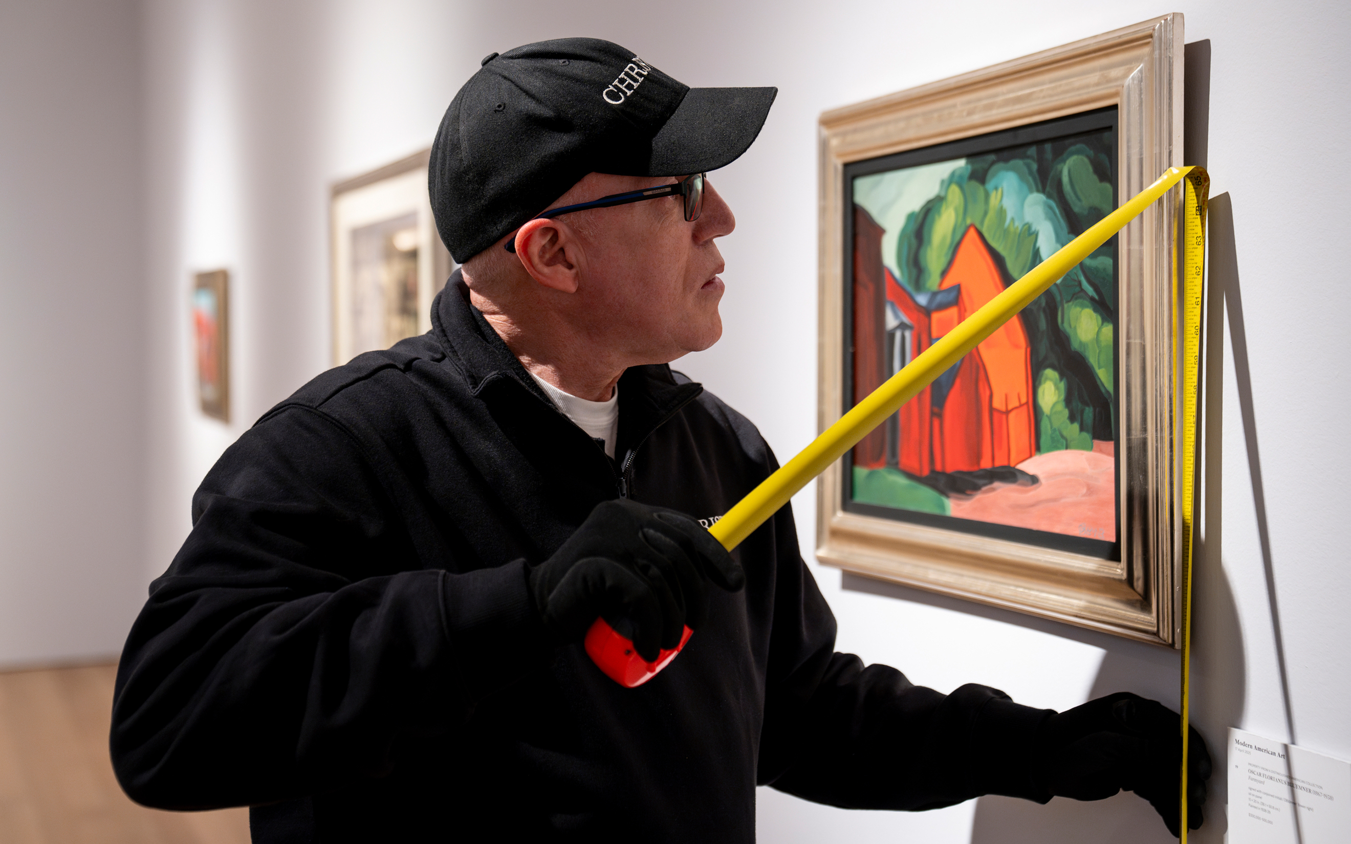 A person in black apparel measures a colorful framed painting in an art gallery using a yellow tape measure.
