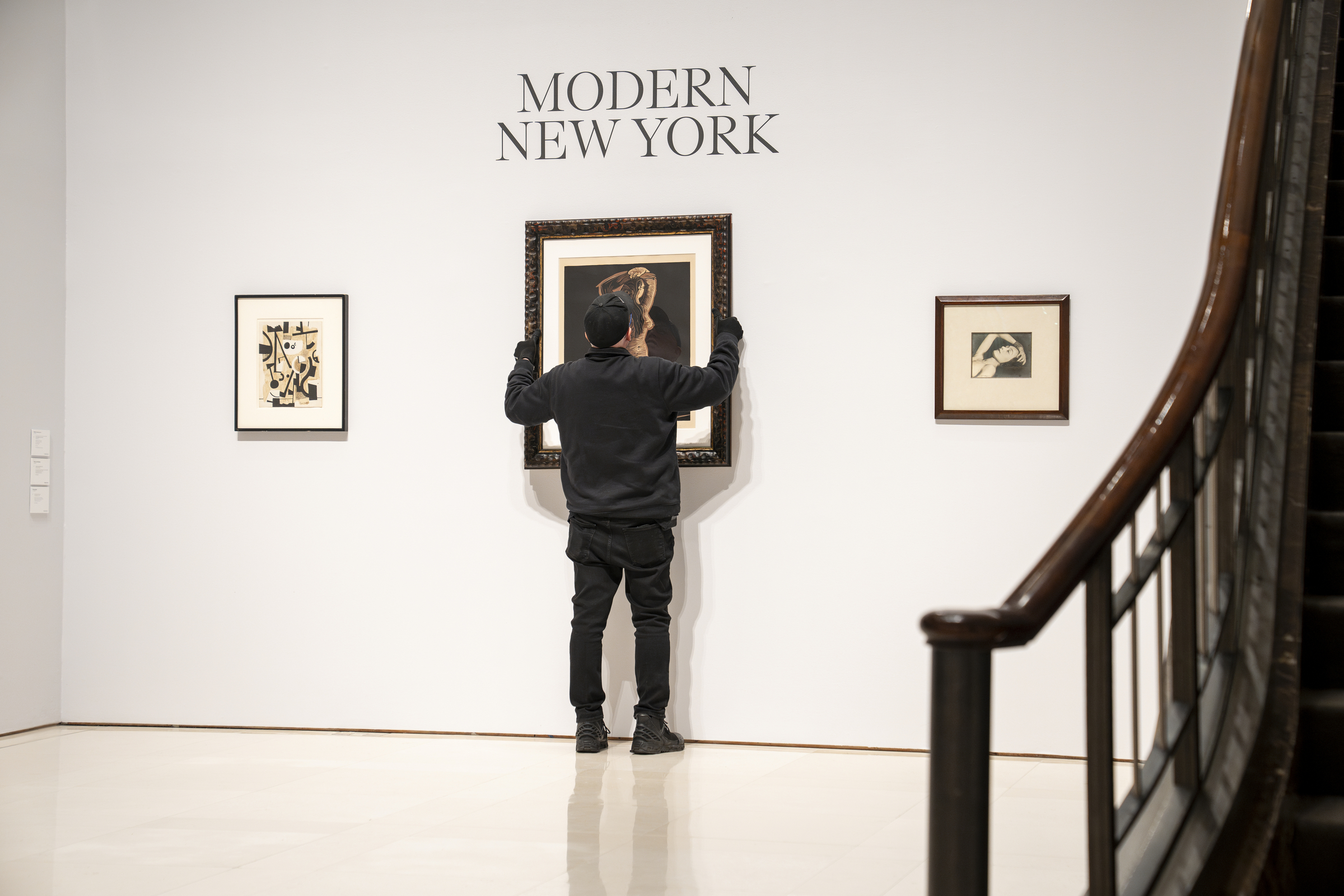 A person in black adjusts art in a gallery labelled "MODERN NEW YORK," next to a staircase.