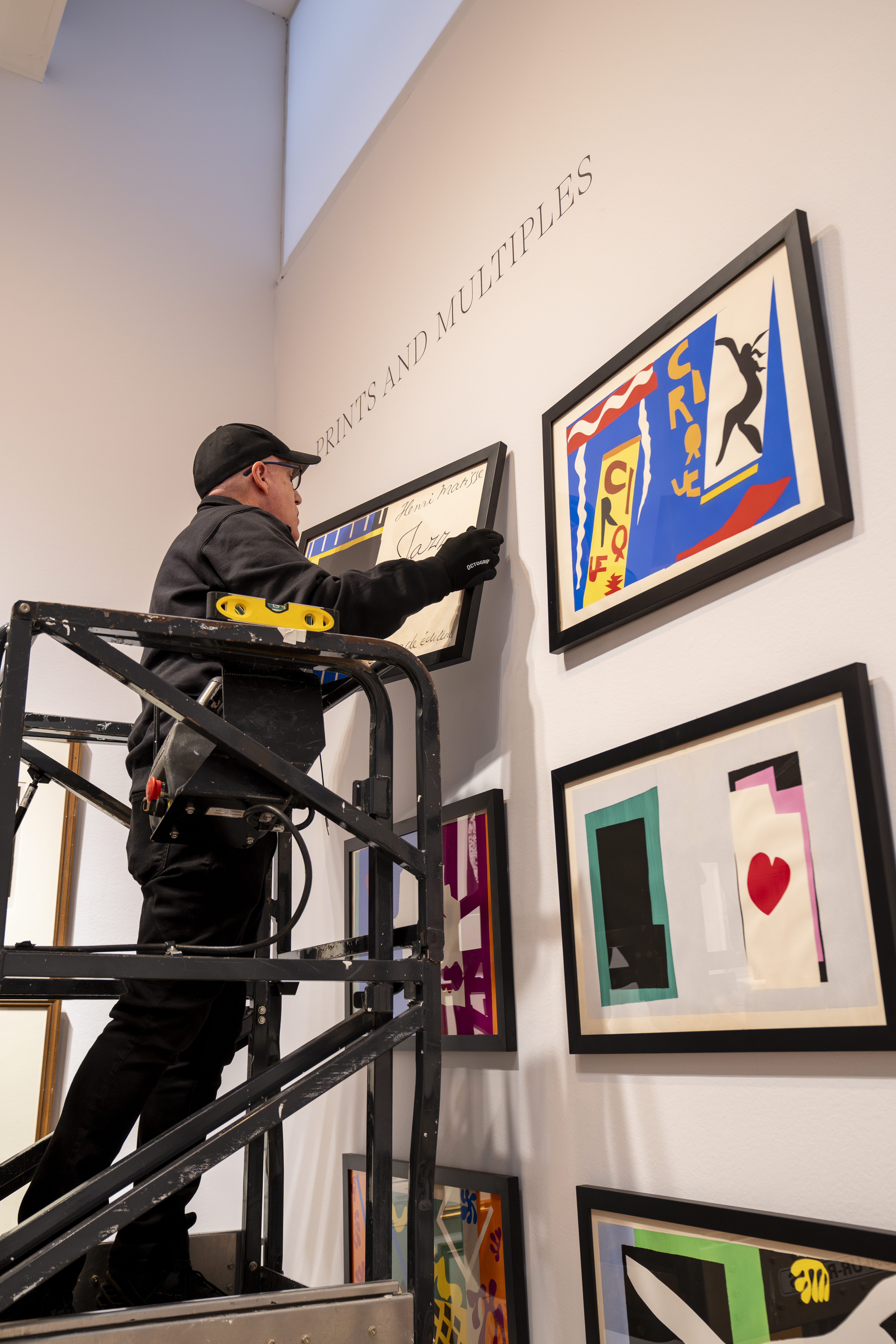 A man on a scissor lift adjusts a framed artwork in a gallery with multiple colorful prints on the wall.