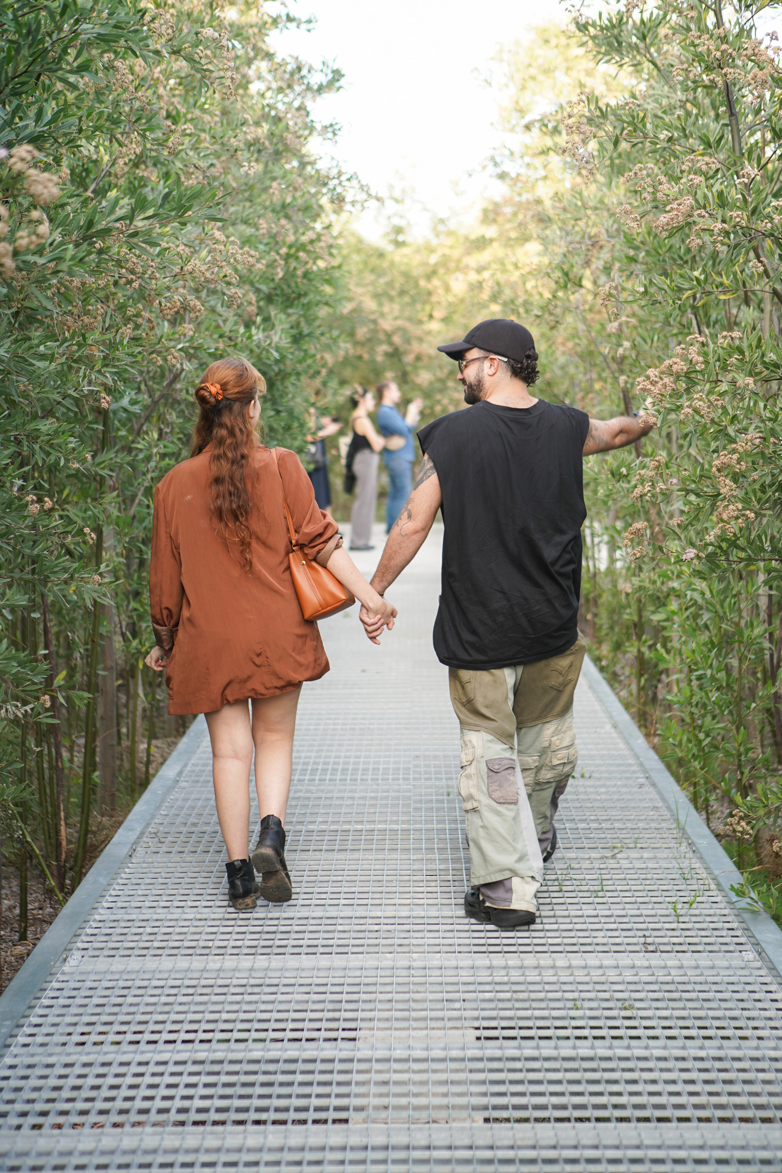 A couple holds hands while walking down a metallic pathway lined with lush greenery.