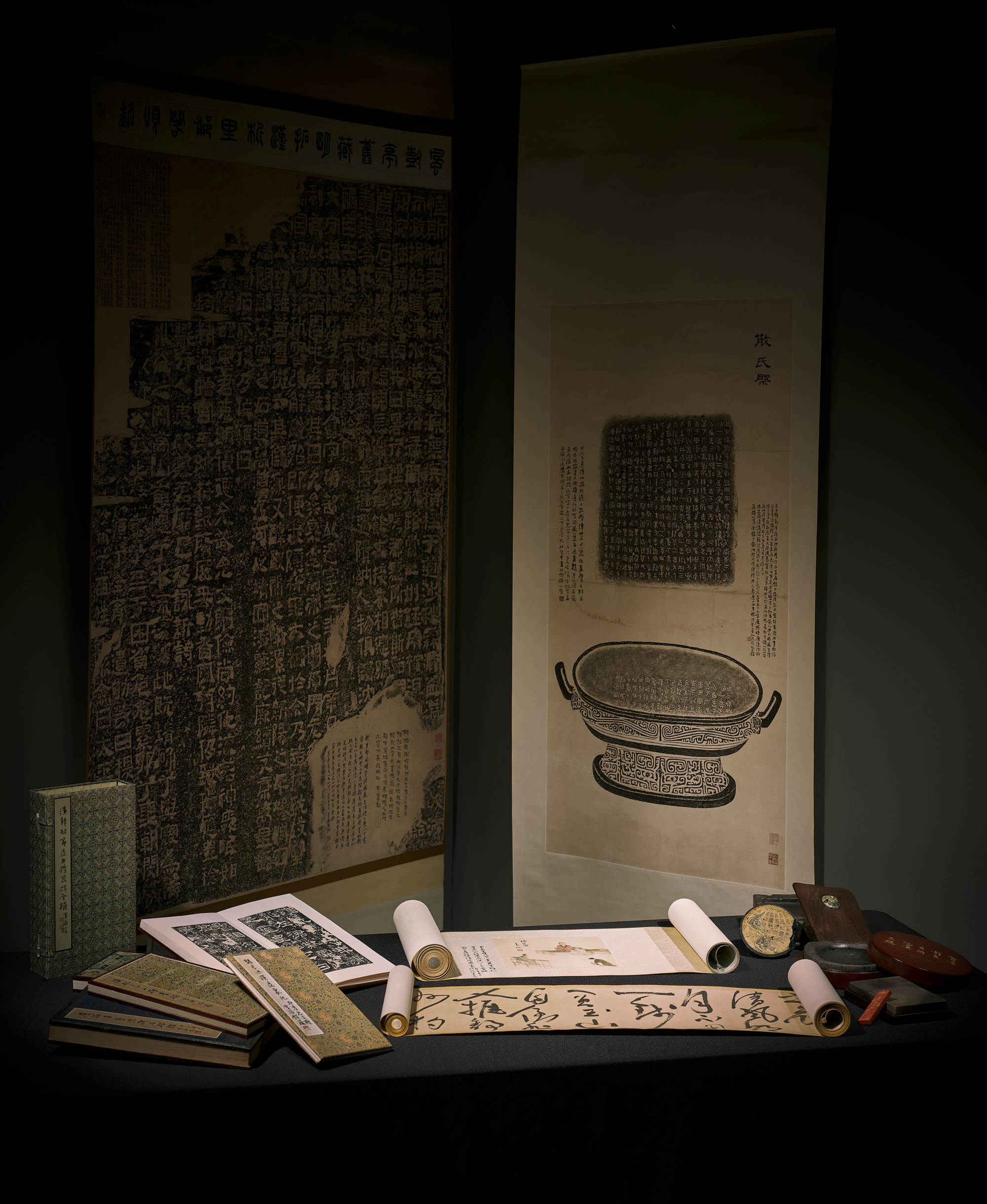 Historical scrolls, books, and artifacts, displaying Chinese text and illustrations, arranged meticulously on a table and against a backdrop.