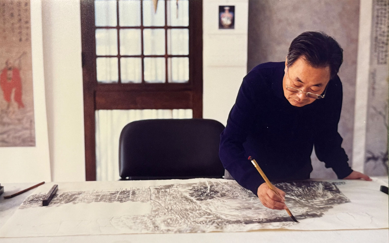 An individual is working on a detailed drawing at a desk adorned with artistic tools in a classically styled room.