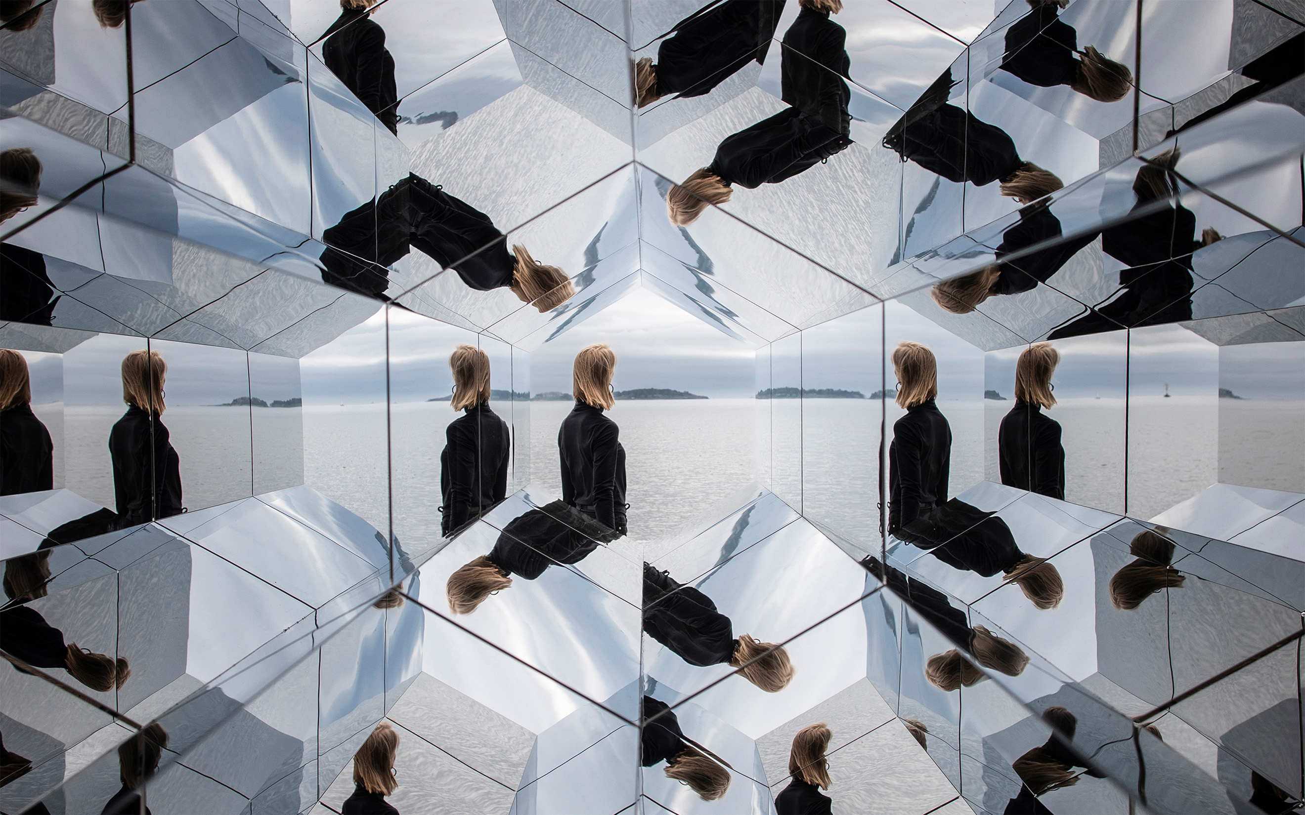 A woman appears multiple times in reflections created by an array of mirrored tiles, set against a serene lake backdrop.