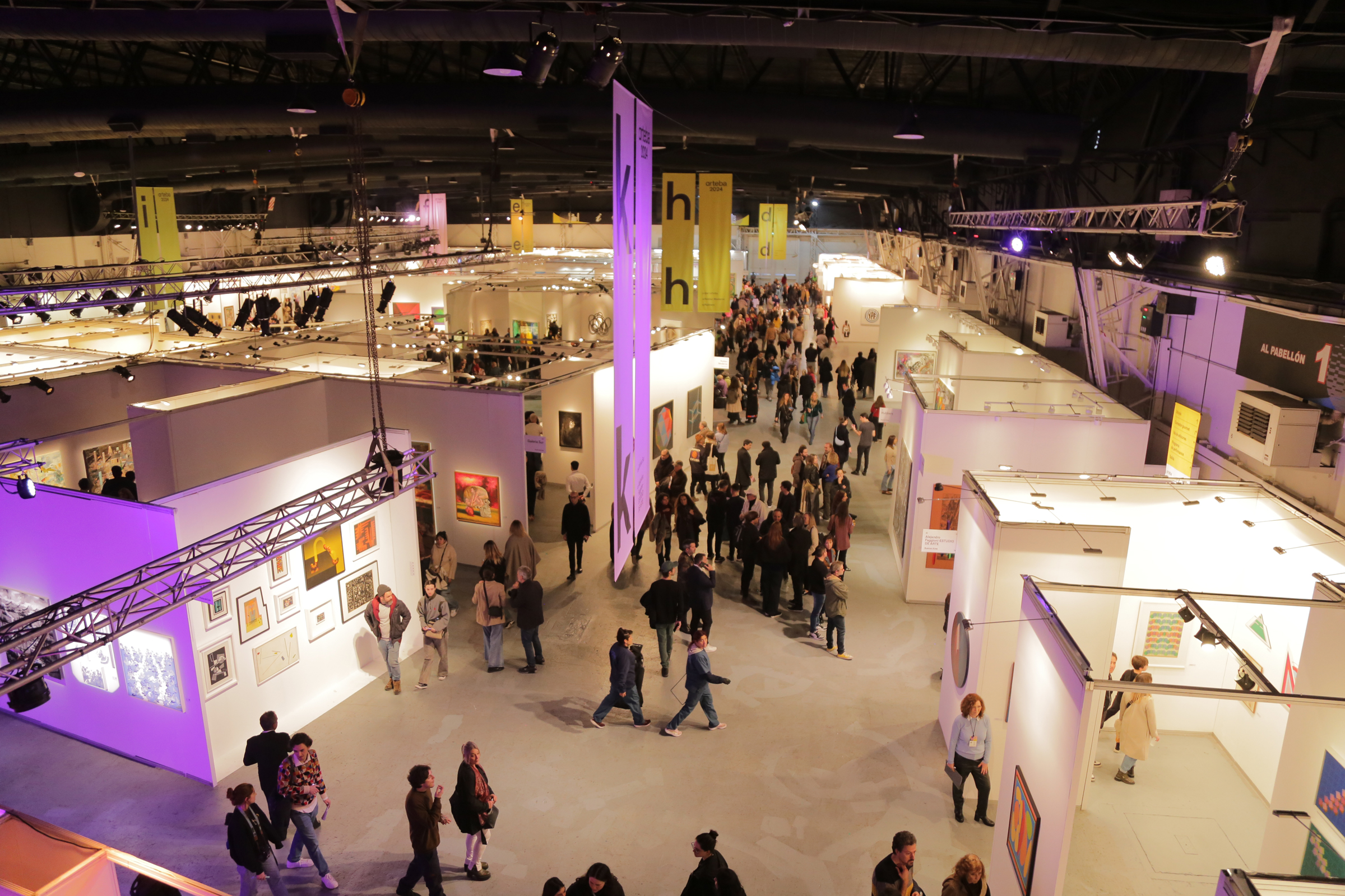 An expansive indoor art fair featuring numerous booths displaying various artworks, with visitors walking around and examining the pieces.