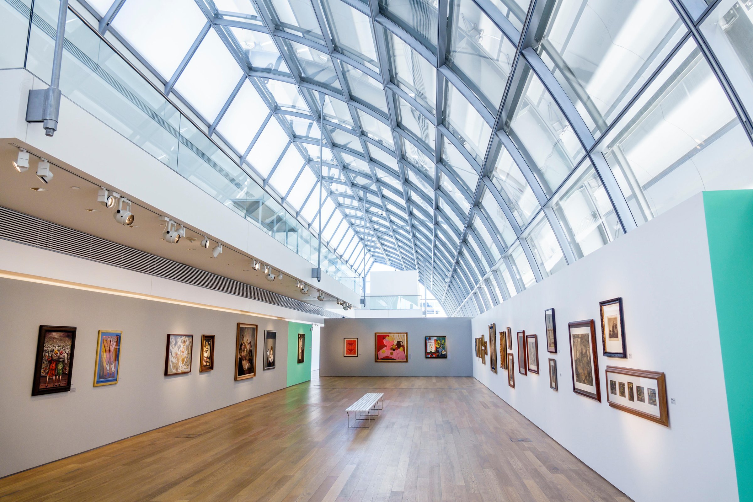 A modern art gallery with a glass ceiling and colorful paintings displayed on white walls.