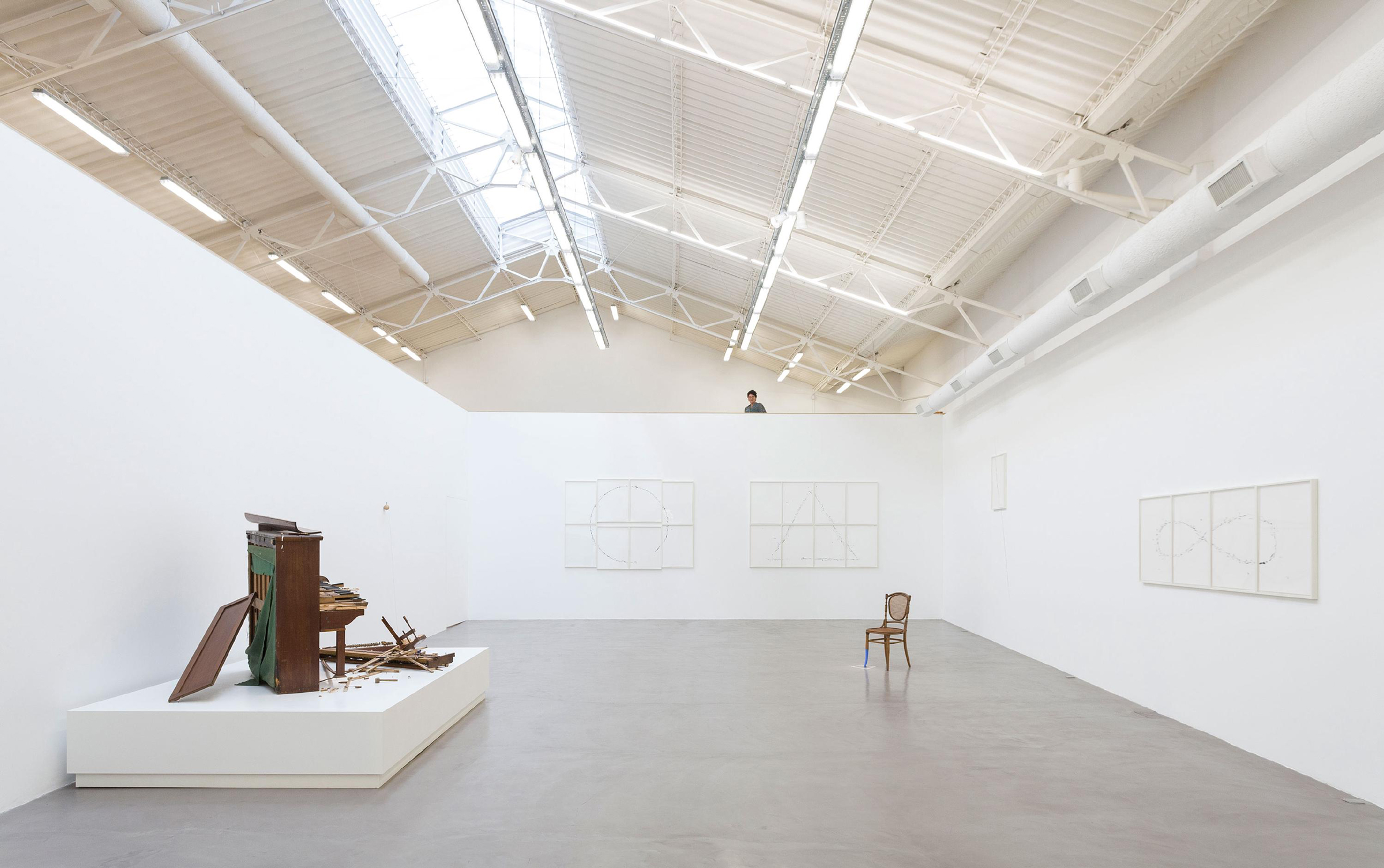 The image shows a spacious art gallery with white walls, displaying various artworks including a large sculptural piece and framed drawings.