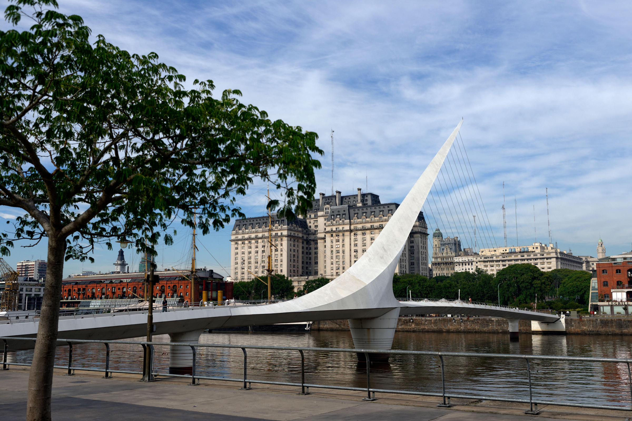Modern pedestrian bridge with a striking white arch contrasts with historic riverside architecture under a bright sky.