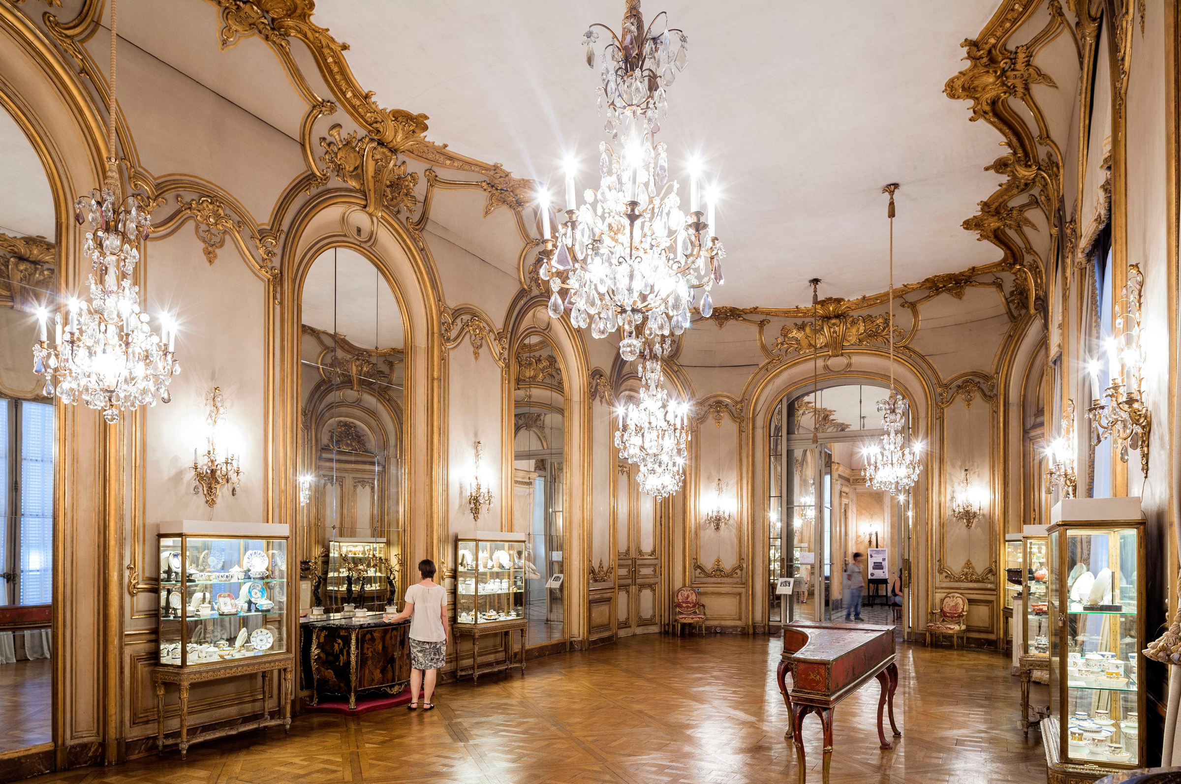 Elaborate room with ornate gold trimmings and multiple chandeliers, featuring display cases and a person viewing items.