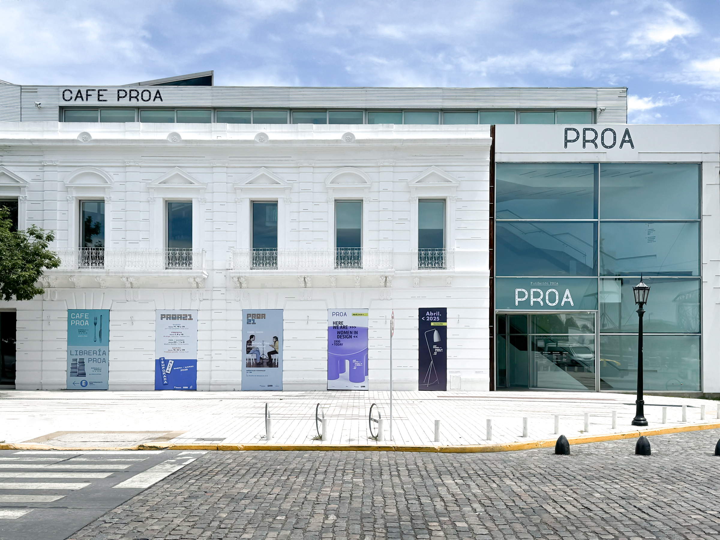 Modern white building with large windows labeled "PROA," featuring an area named "CAFÉ PROA" under a clear sky.
