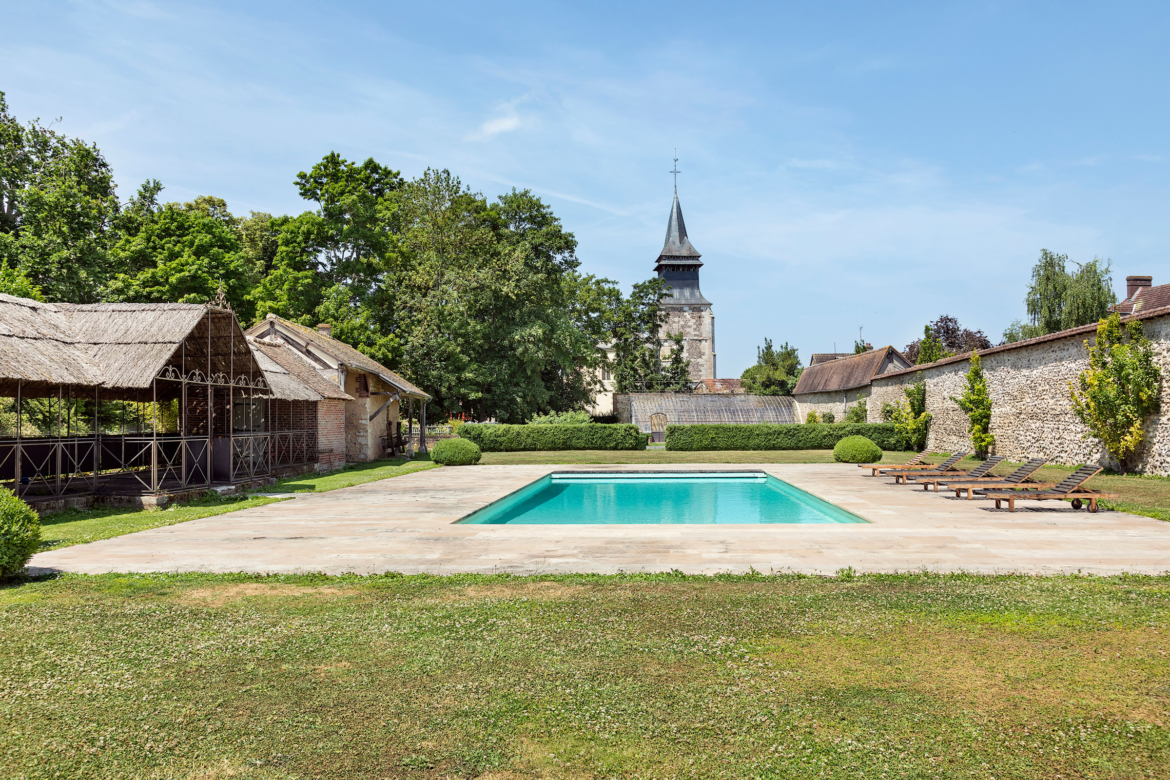 A tranquil pool area with lounge chairs, surrounded by historic buildings and lush greenery under a clear blue sky.