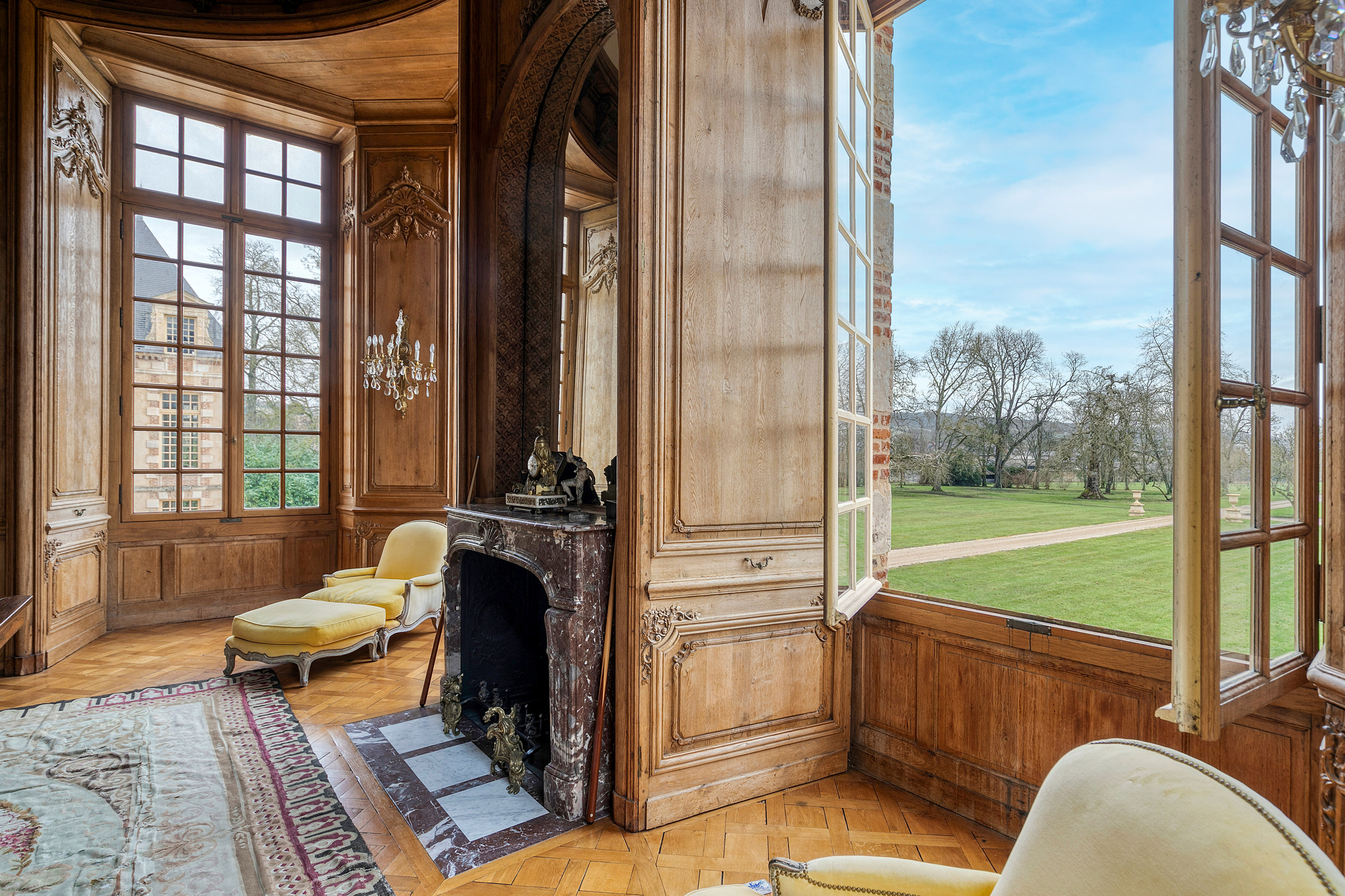 An elegant, richly paneled room with a classic fireplace and large window revealing a park view.