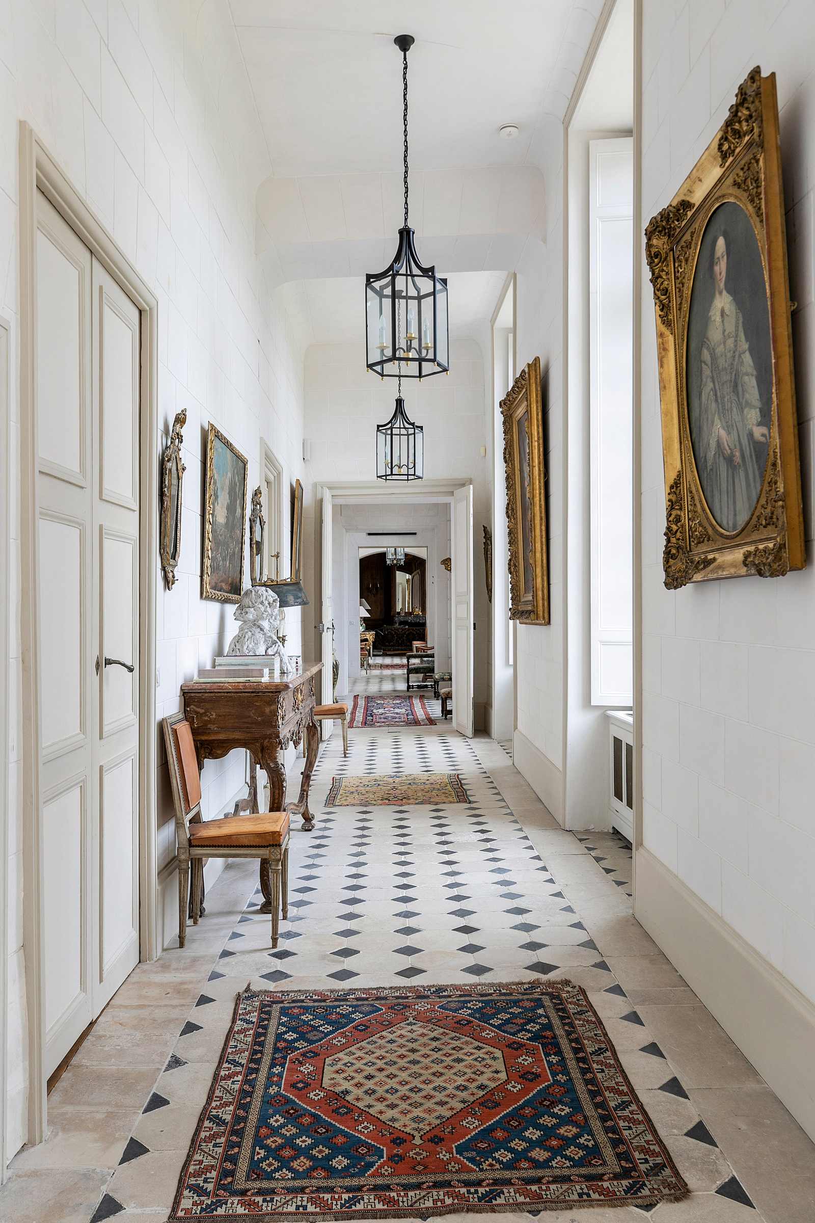 Elegant hallway with black and white tiled floor, adorned with vintage portraits, a hanging lantern, and colorful rugs.