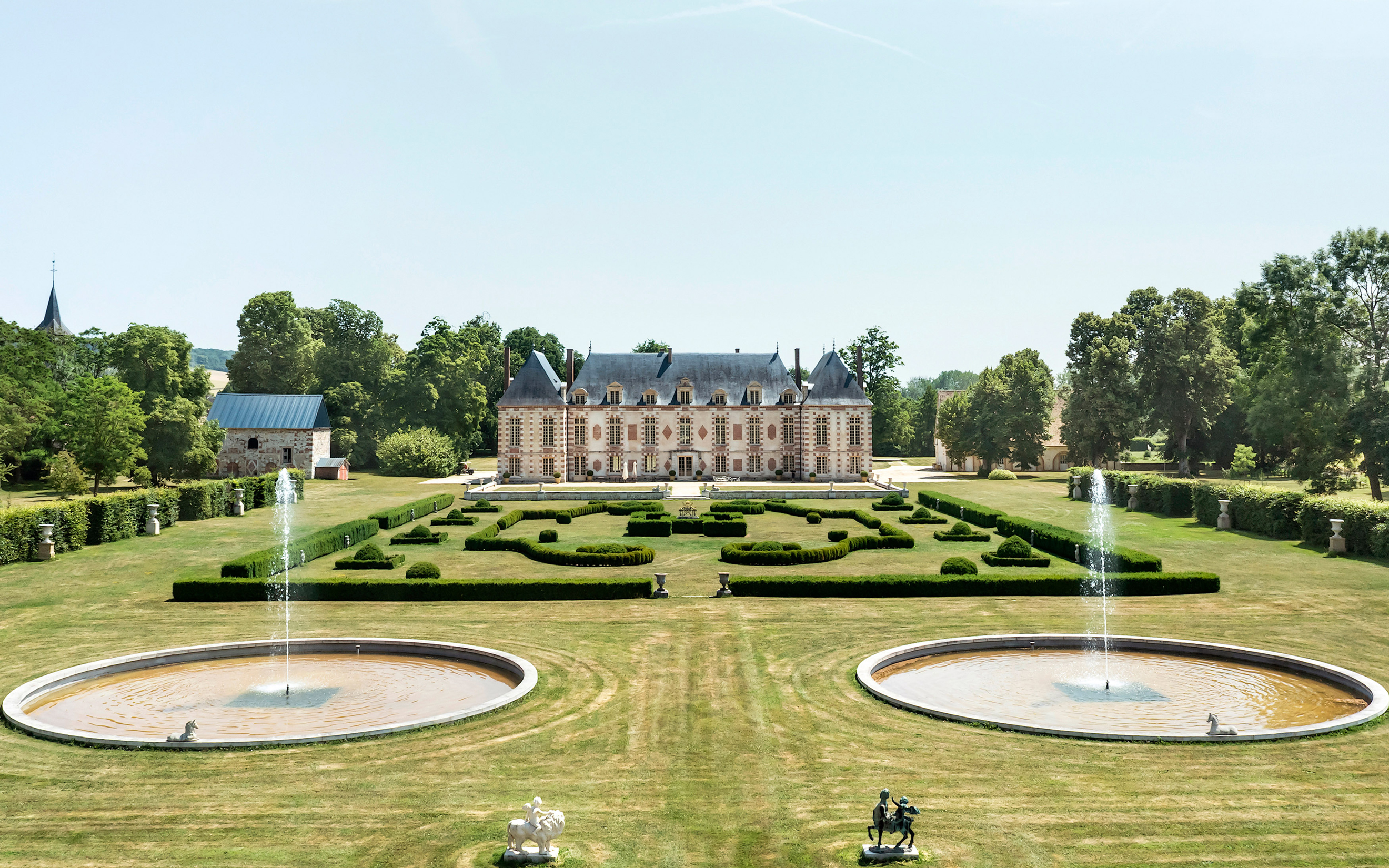 An elegant estate with symmetrically designed gardens and multiple fountains, surrounded by lush trees.