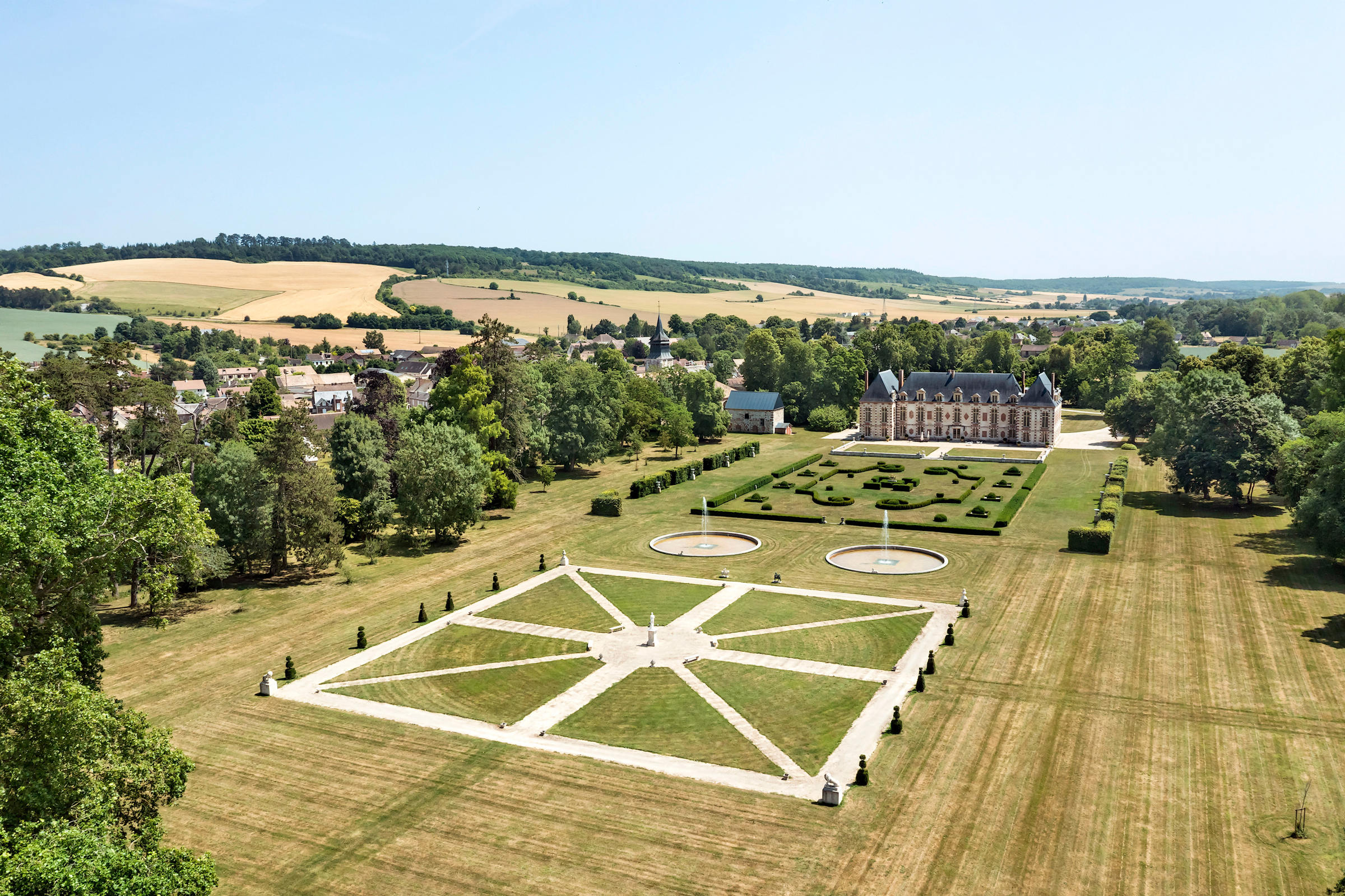 Aerial view of a grand chateau with elaborate gardens, surrounded by a lush landscape and a small village.