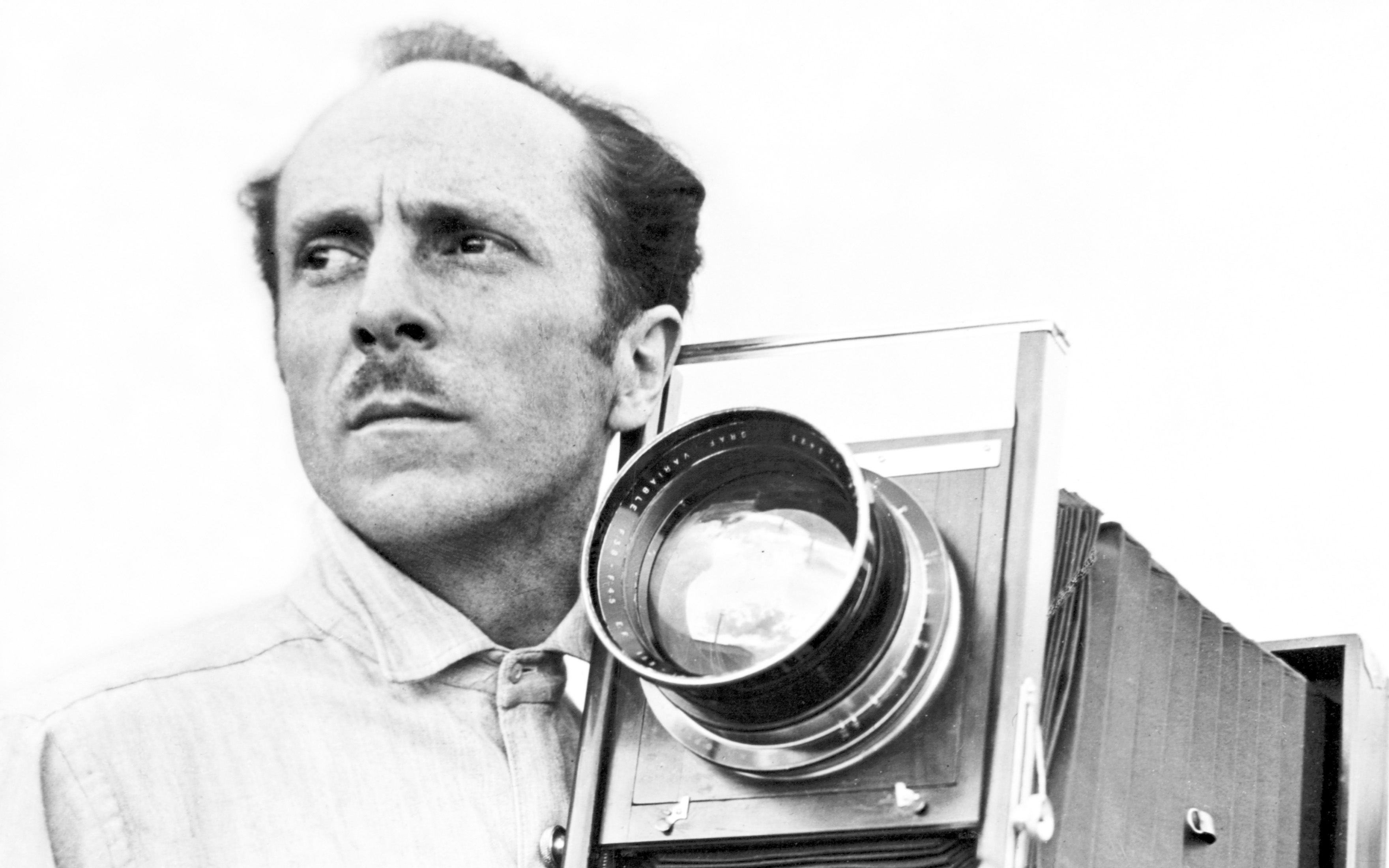 Edward Weston holds a large vintage camera, dressed in a light-colored shirt.