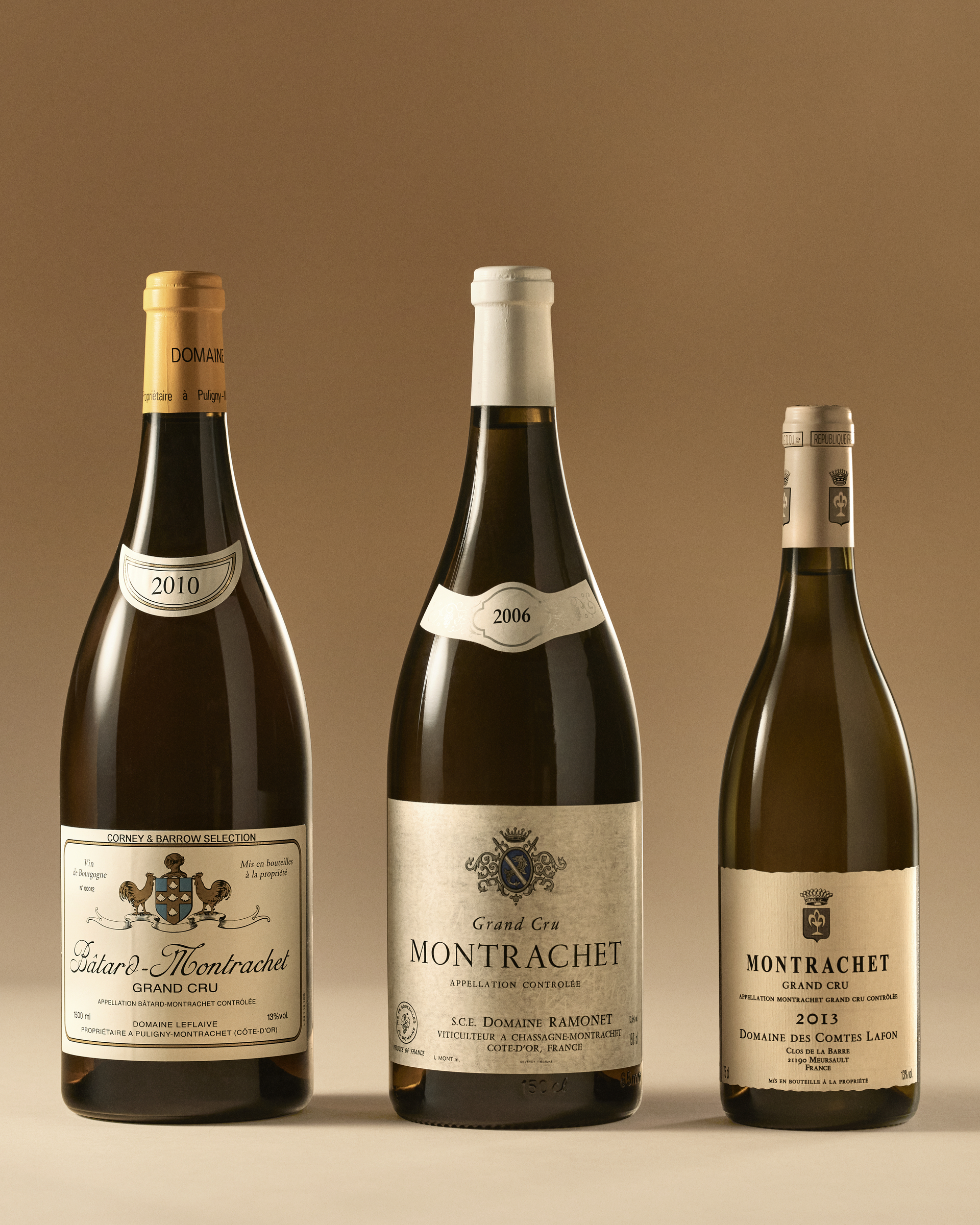 Three bottles of Montrachet Grand Cru white wine from different vintages and producers, displayed against a beige background.