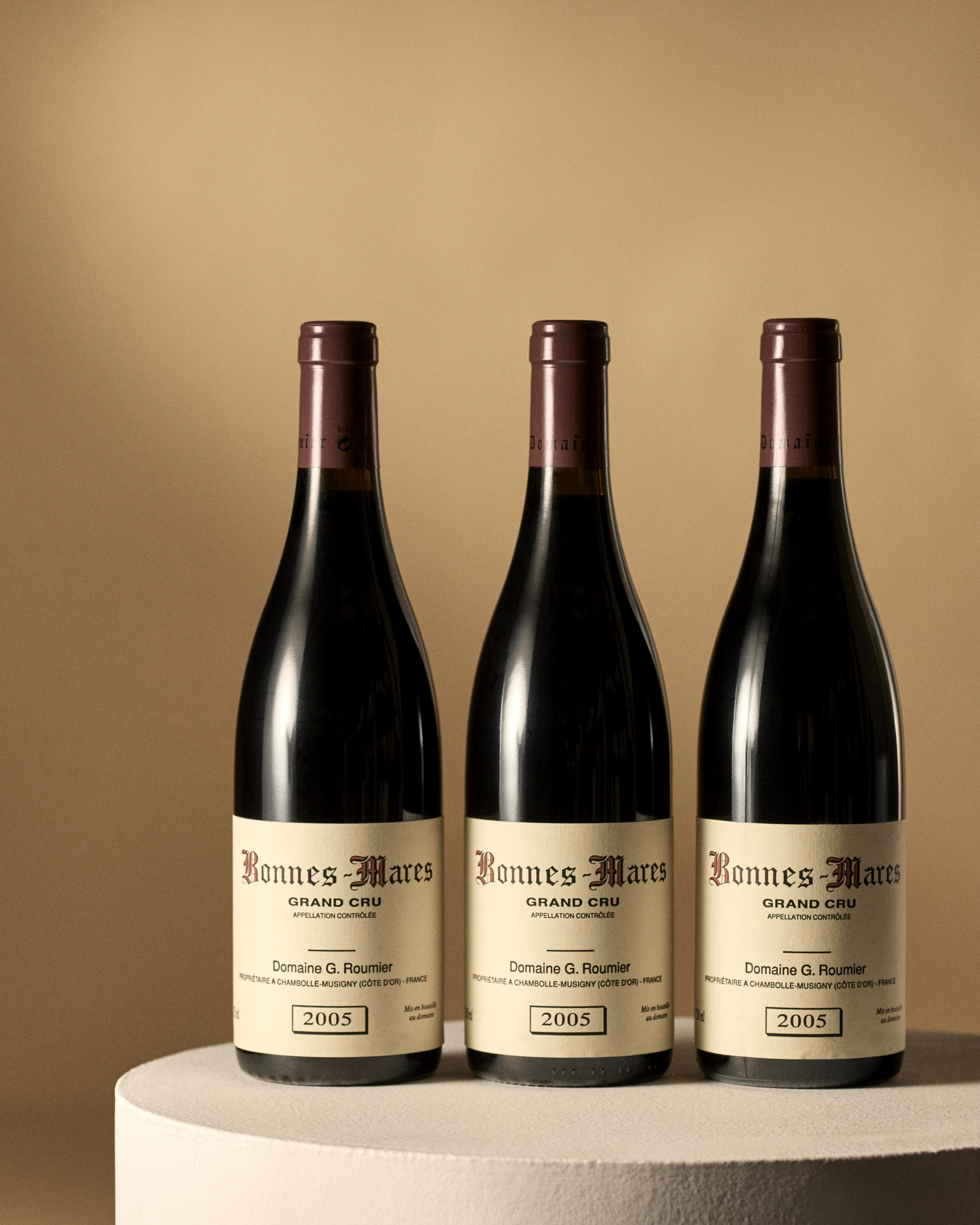 Three bottles of Bonnes-Mares Grand Cru 2005 wine displayed on a neutral-colored platform with a beige background.