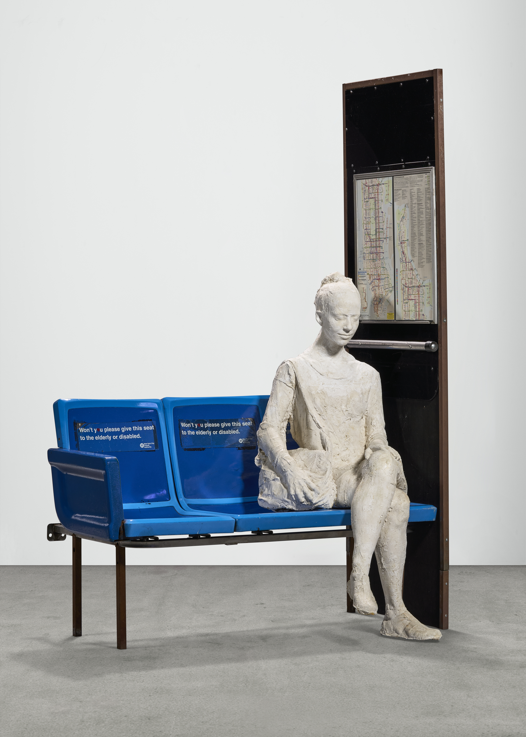 A sculpture sits on blue public transit seats next to an open, map-lined cabinet, creating a scene reminiscent of a subway station.