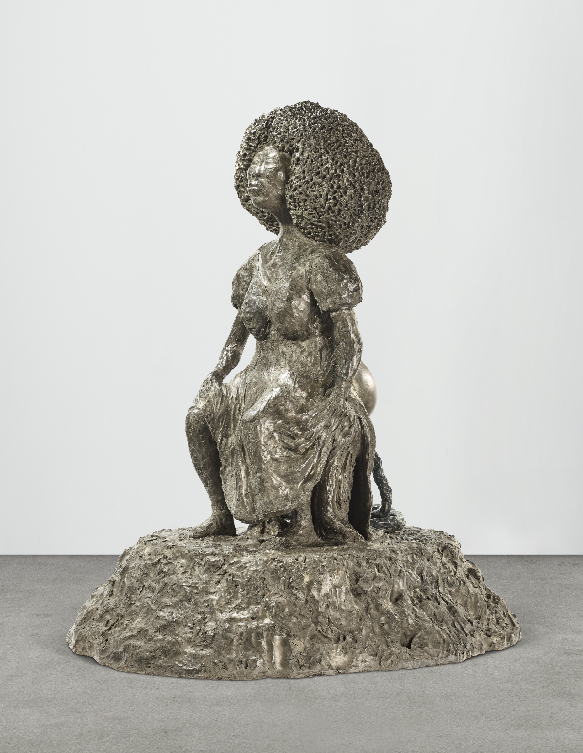 A sculpture of a seated female figure with an elaborately textured, voluminous hairdo, rendered in bronze.