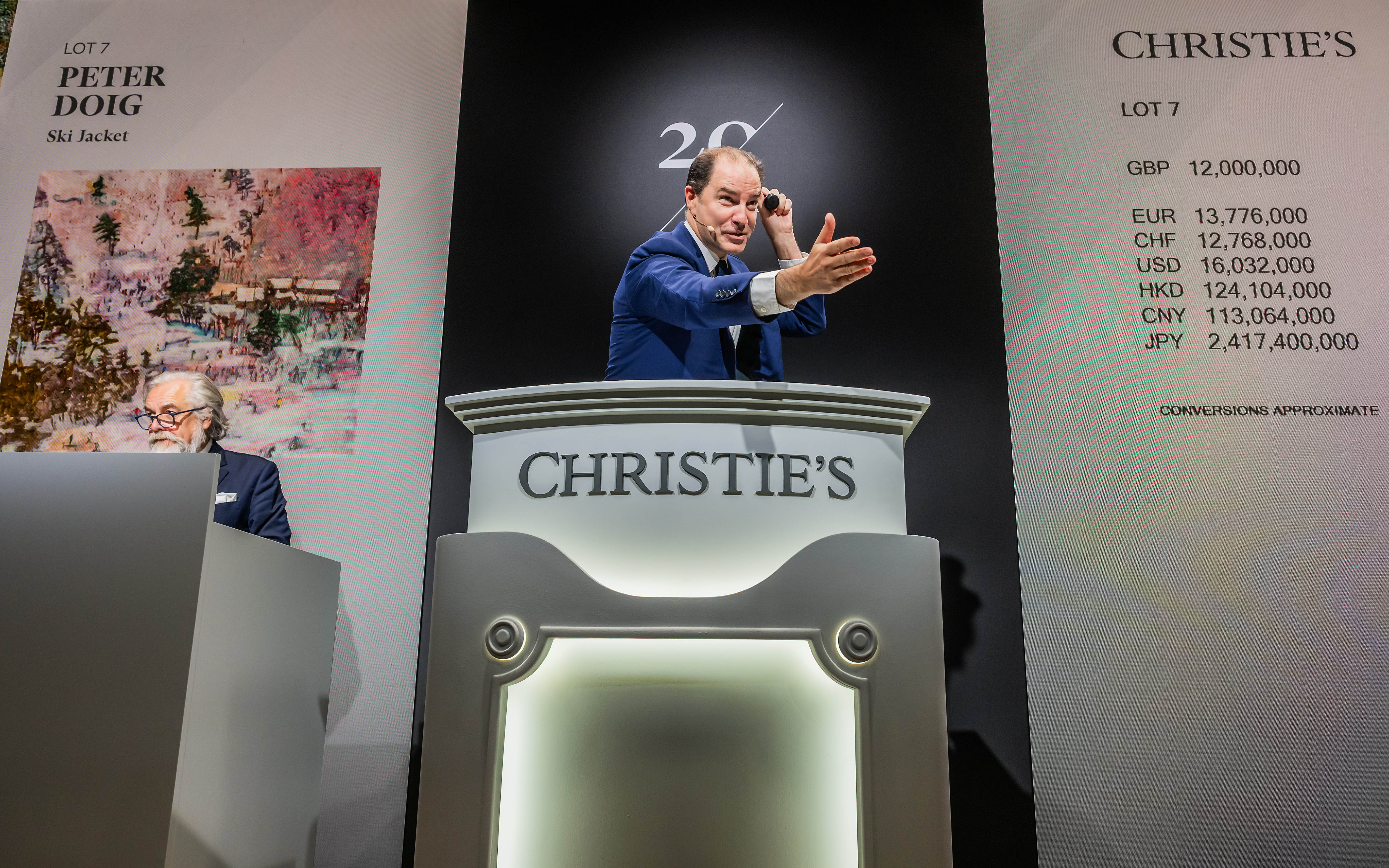 The image shows an auction at Christie's with a man gesturing near a podium with currency conversions listed for a Peter Doig painting titled "Ski Jacket."