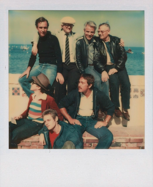 A group of individuals in various casual and semiformal outfits sits on a ledge by the sea, posing in a relaxed manner.