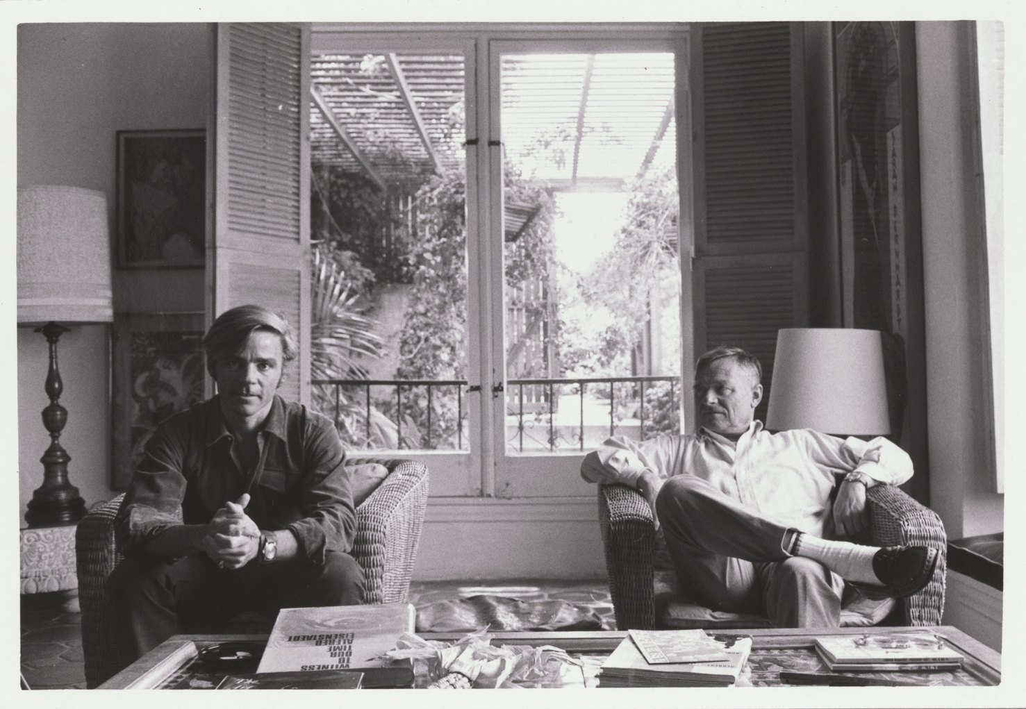 Two individuals are sitting in a living room with wicker chairs, a coffee table covered with various items, large windows, and a visible garden outside.