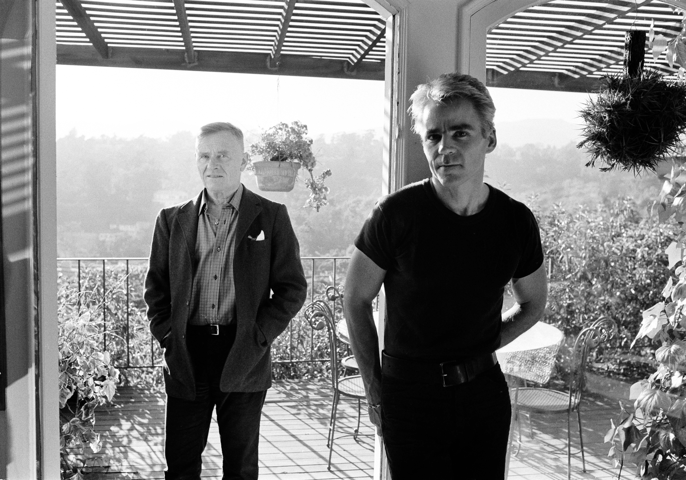 Two men stand on a balcony surrounded by plants and overlooking a lush landscape, dressed in contrasting formal and casual outfits.