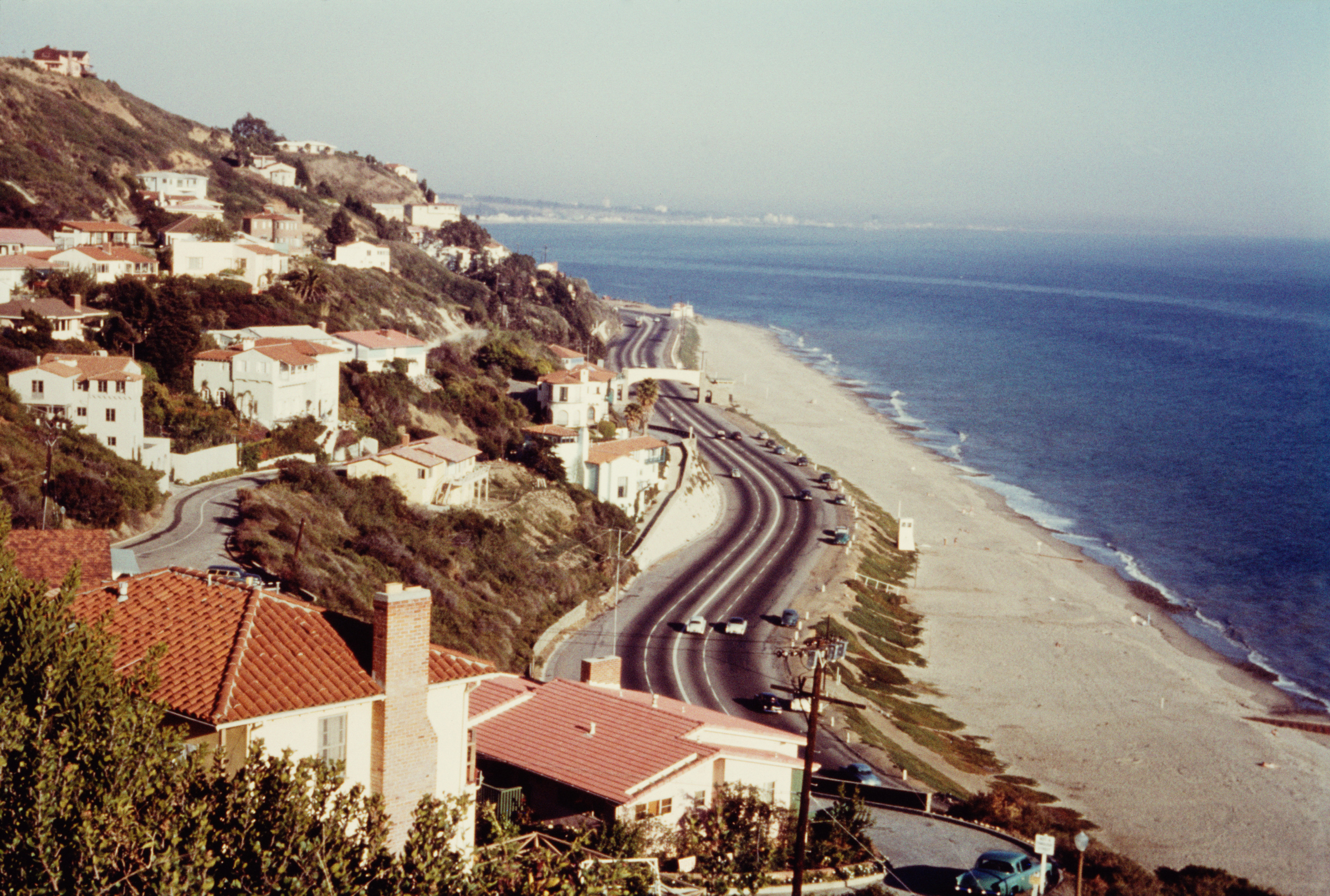 A coastal road winding past beachfront and hillside homes, overlooking a vast, sandy beach and ocean.