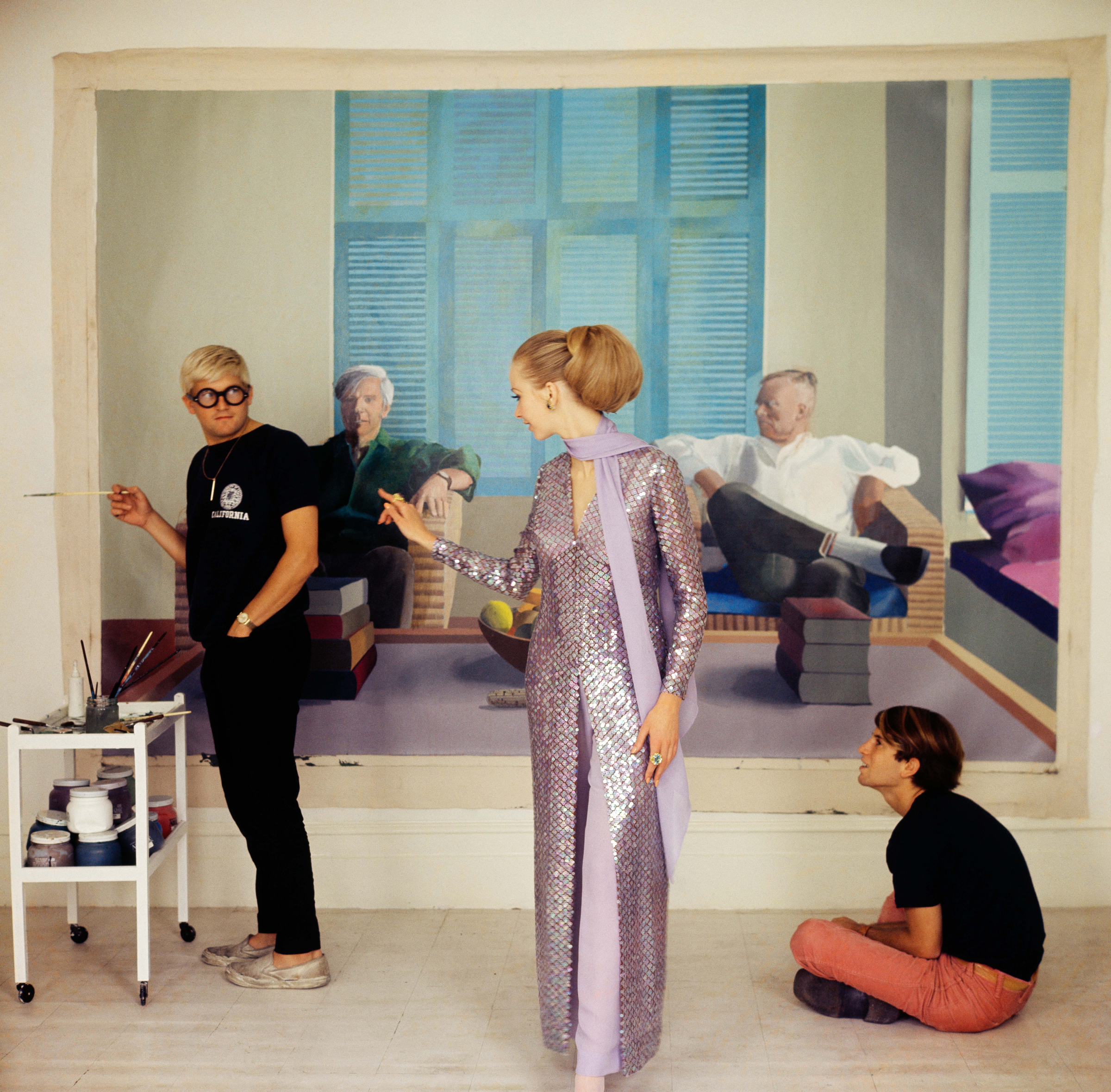 Two individuals are interacting near a painting of people in a living room, with one person sitting on the floor and one holding a paintbrush.