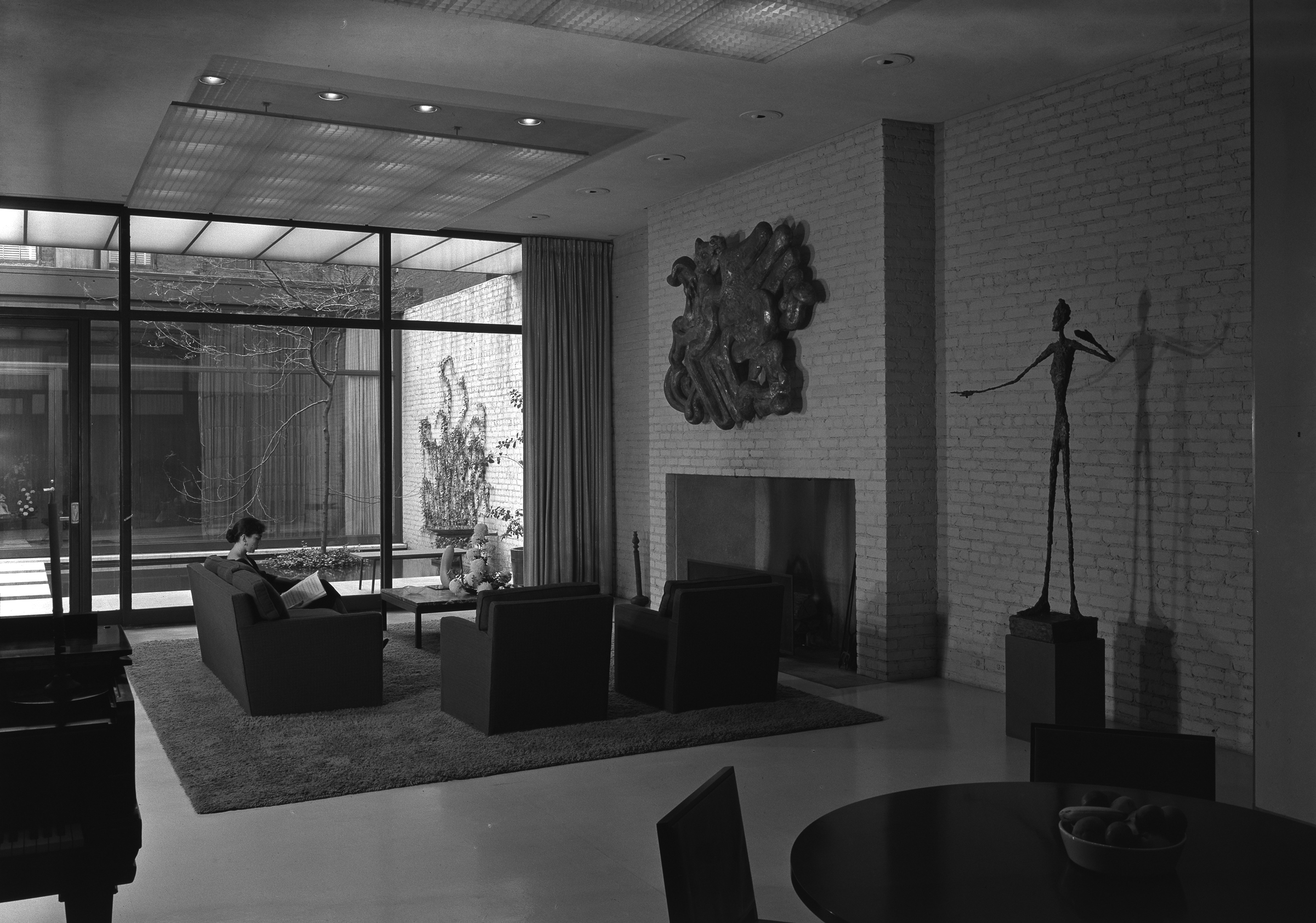 A woman reads in a stylish, modern living room with large windows, sculptures, and a grand piano.