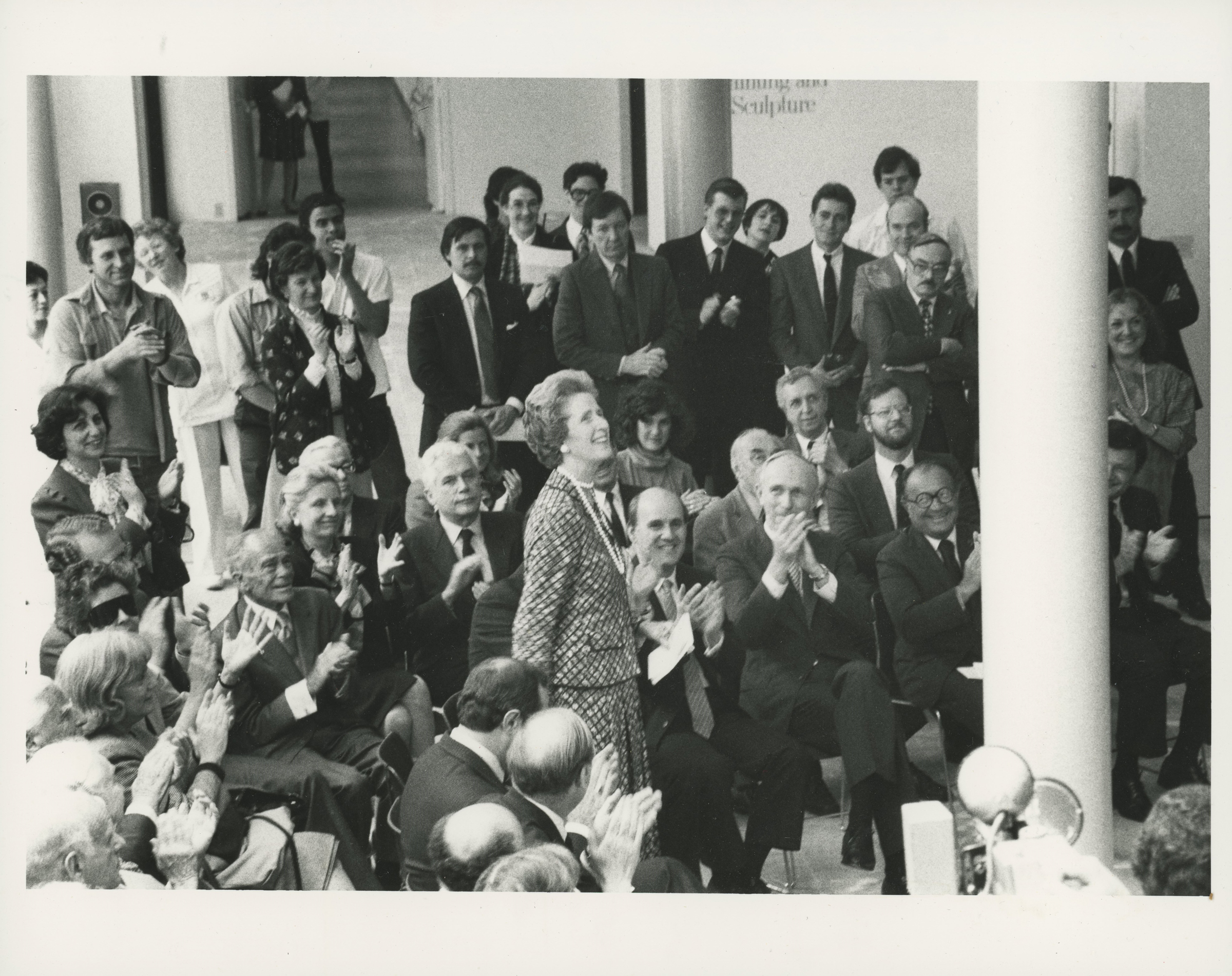 A group of people in formal and casual attire is gathered in a room, clapping, with a sign reading "sculpture" in the background.
