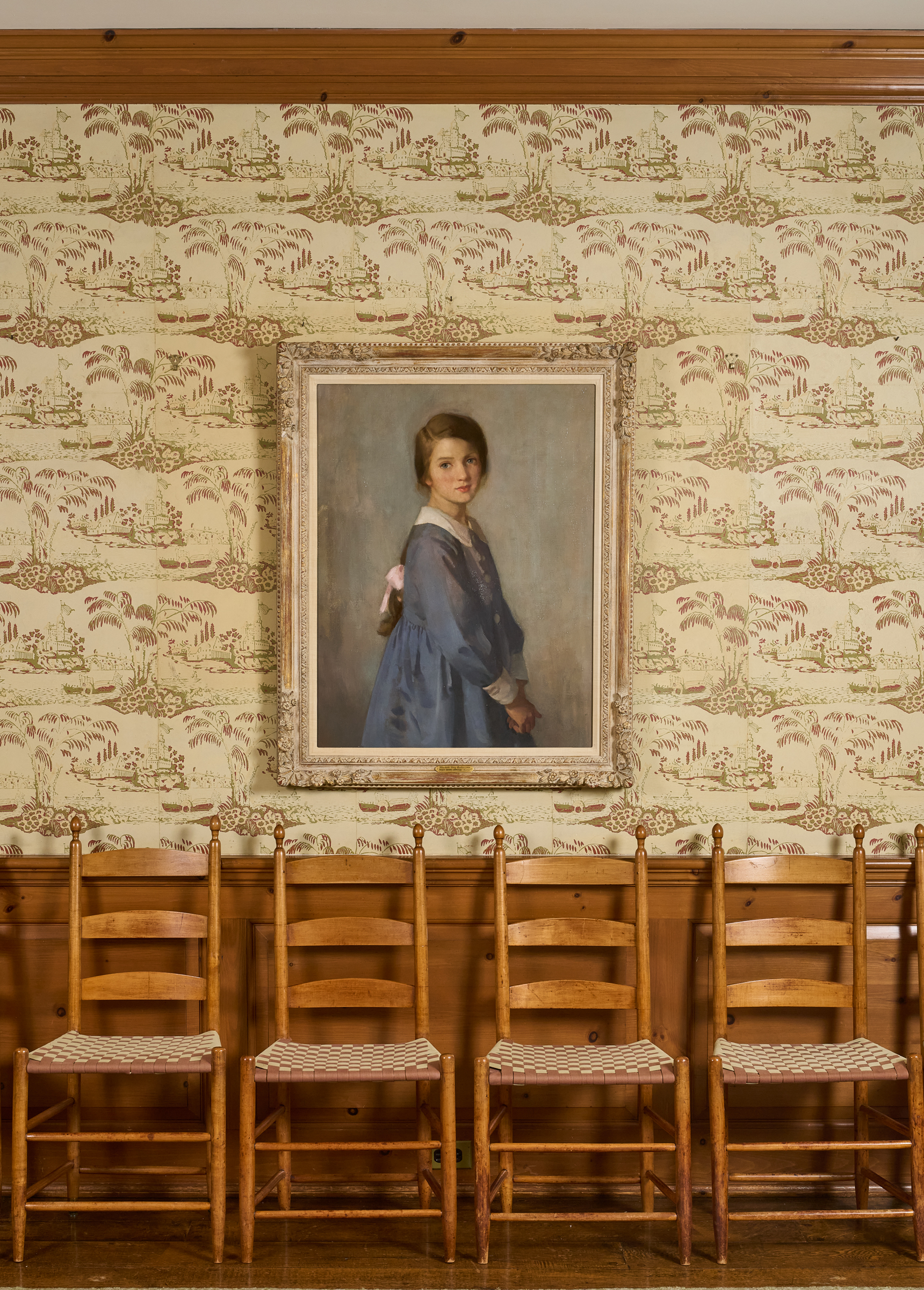 A row of wooden chairs lined against a wall, under a framed painting hung on a patterned wallpaper.