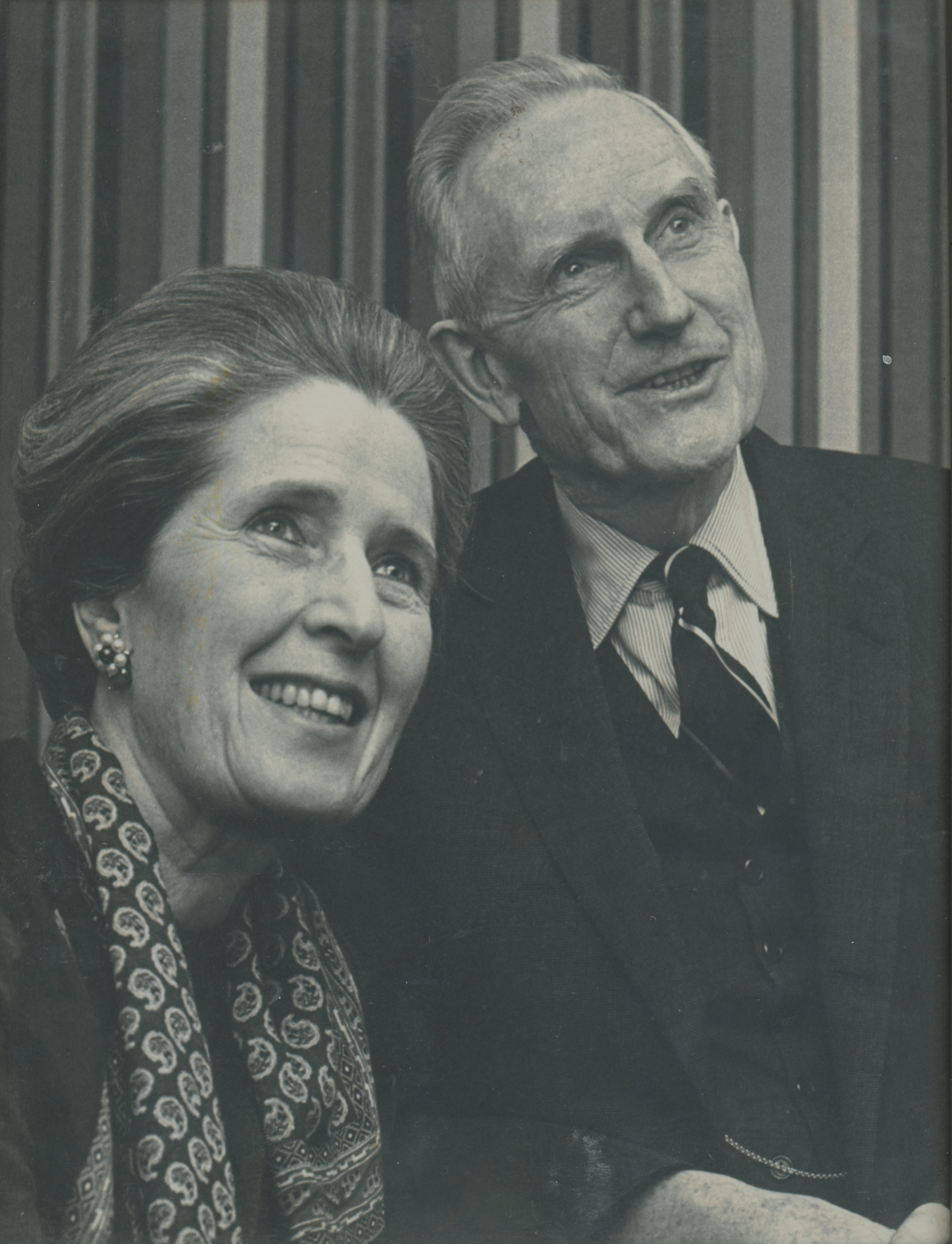 The image shows two people sitting side by side, one dressed in a dark blazer and striped tie, and the other adorned in a paisley shawl.