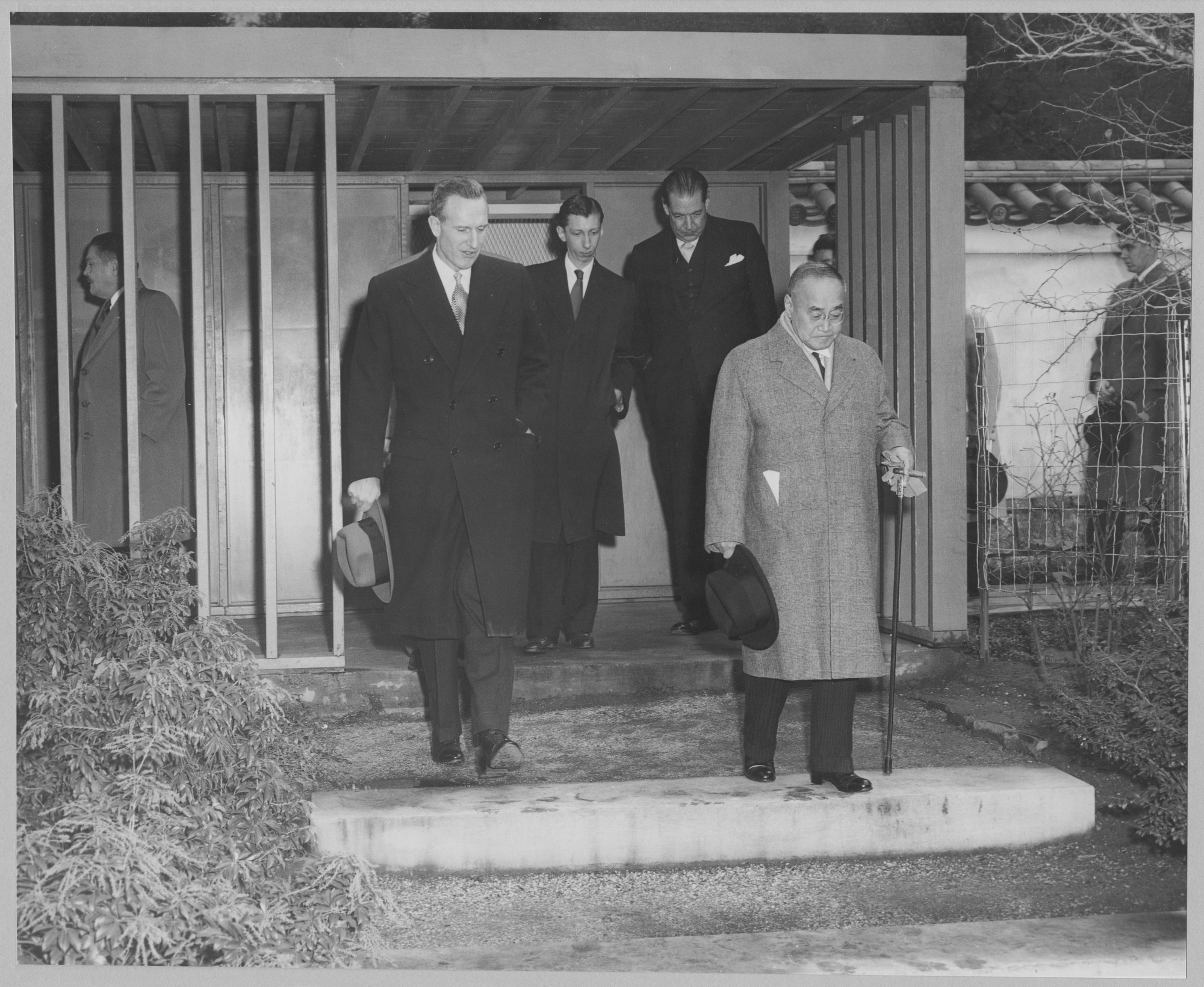 A group of men in formal attire with hats exits a building, walking down steps.
