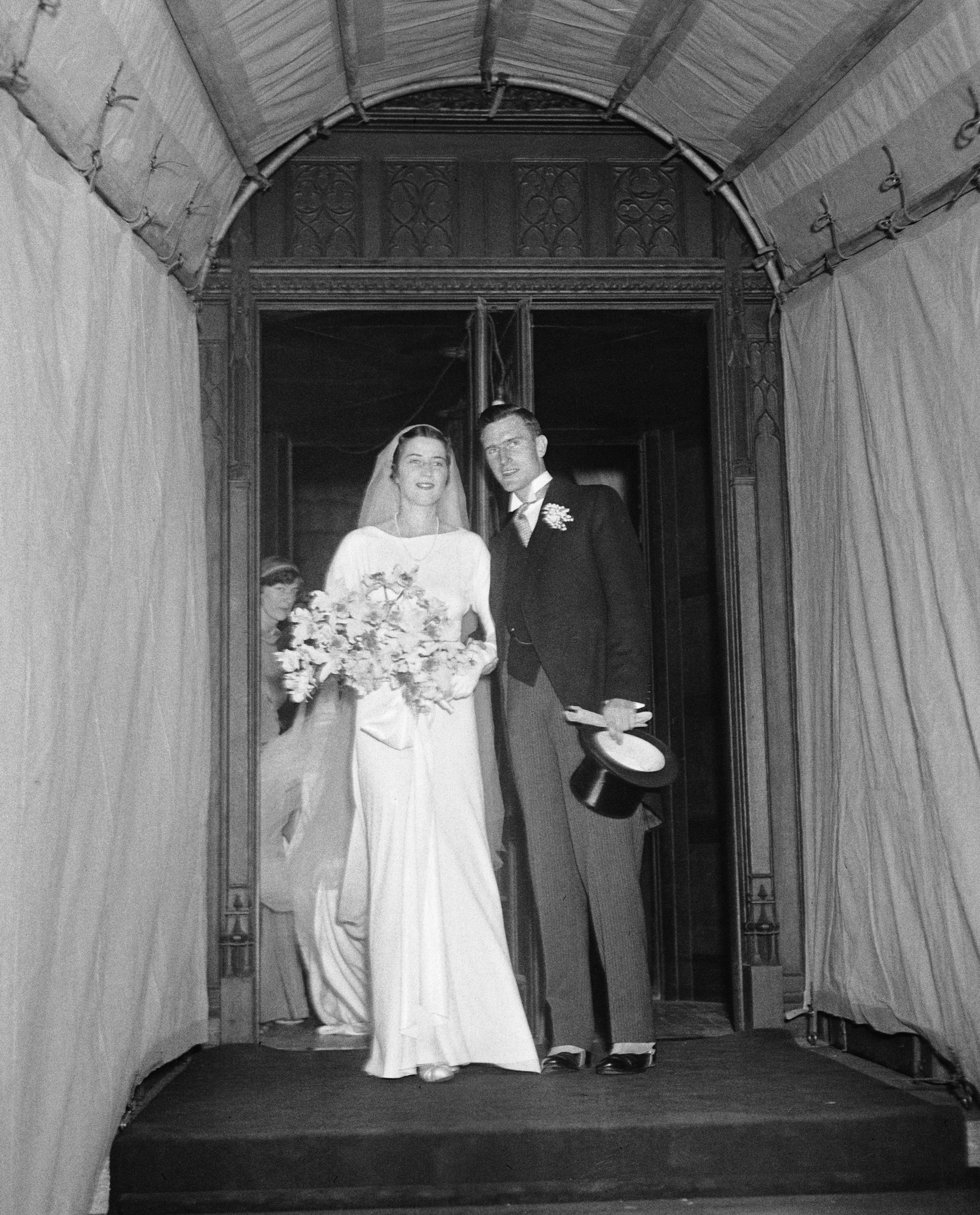 A vintage wedding photo showing a couple exiting a building, the woman in a long white gown with a bouquet and the man in a dark suit and holding a top hat.