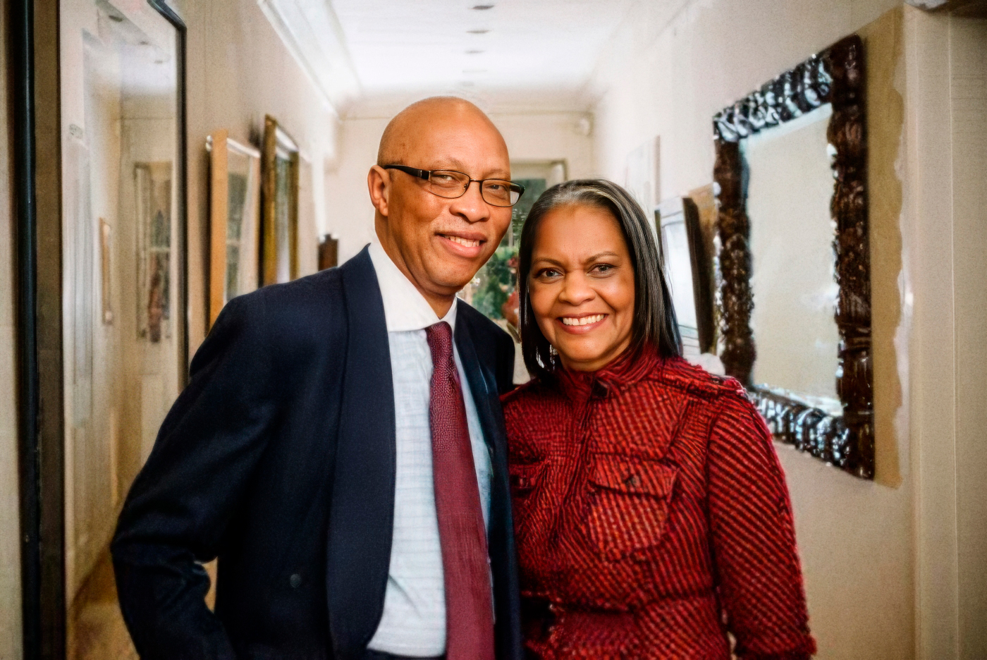 A man in a dark suit and red tie and a woman in a red patterned jacket are standing in a corridor with pictures on the wall.