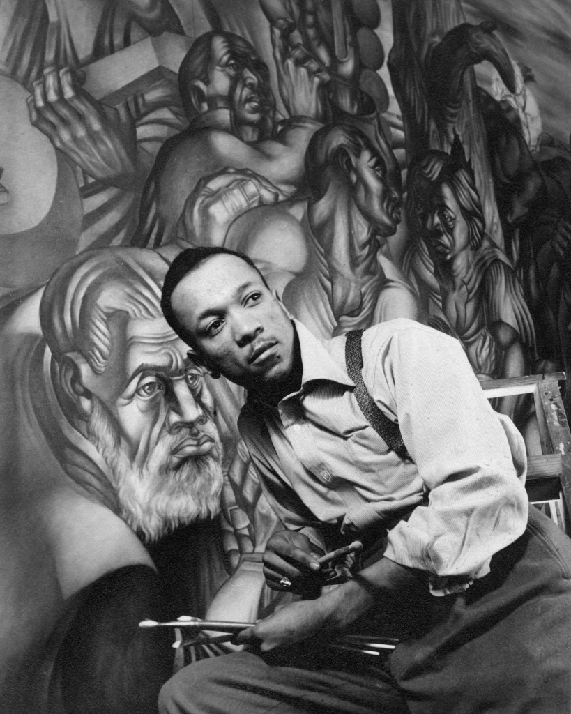 The image shows an artist holding a palette and paintbrushes, sitting in front of a large, detailed mural depicting multiple dynamic human figures.