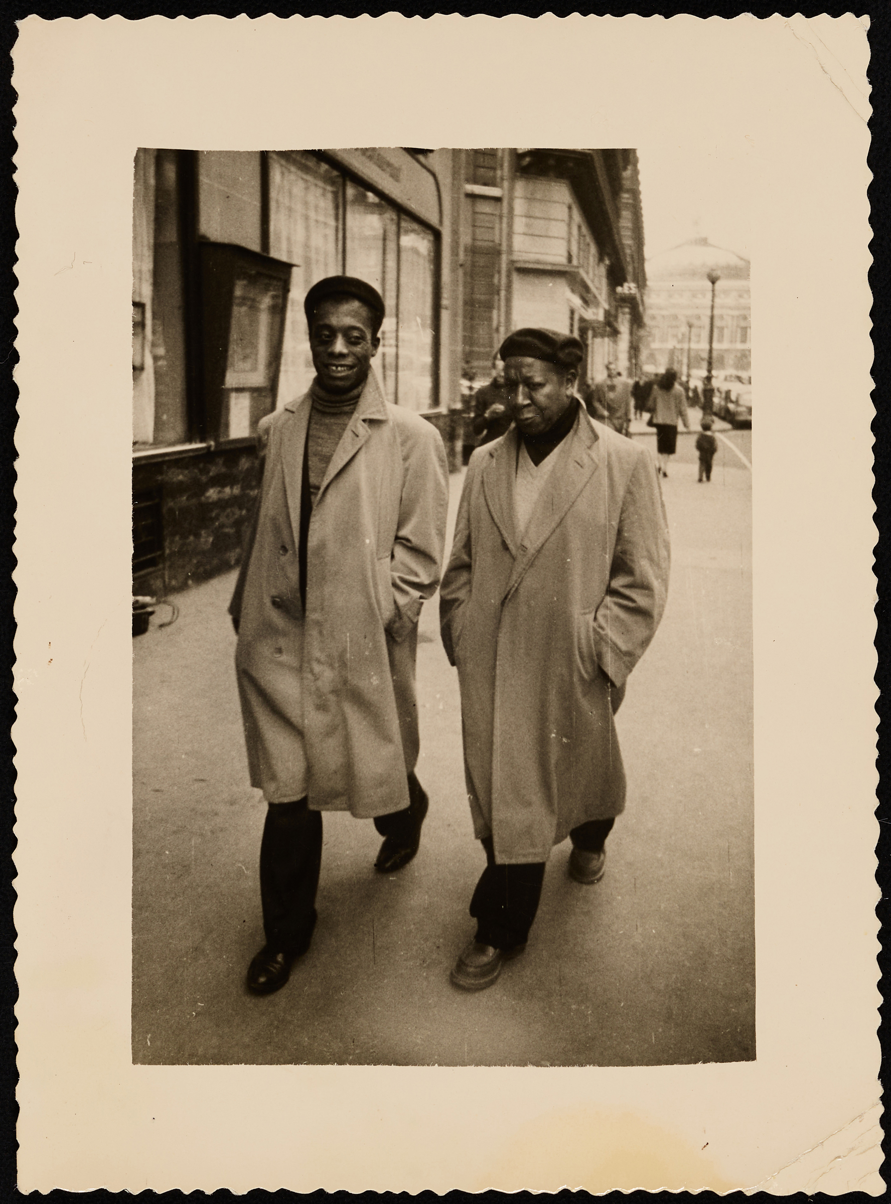 Two individuals in trench coats walk down a bustling city street in a vintage black and white photo.