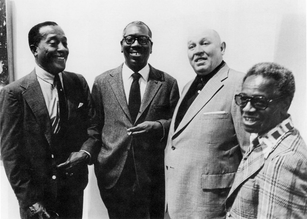 Four men in suits appear in the photograph, standing side by side.