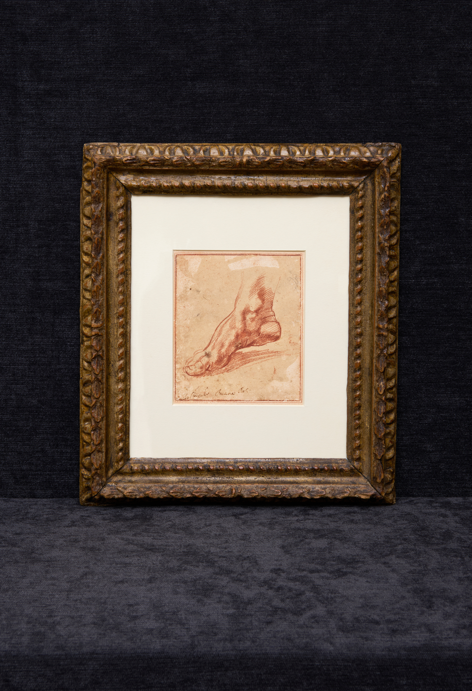 A framed sepia sketch depicts a human foot, displayed against a dark fabric background.