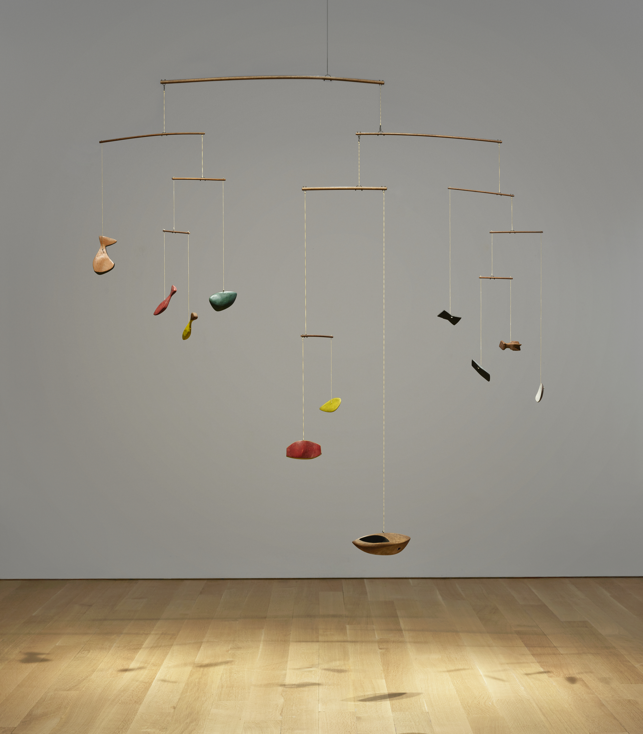 A mobile sculpture with colorful abstract shapes suspended on strings from wooden rods, displayed in a minimalistic room.