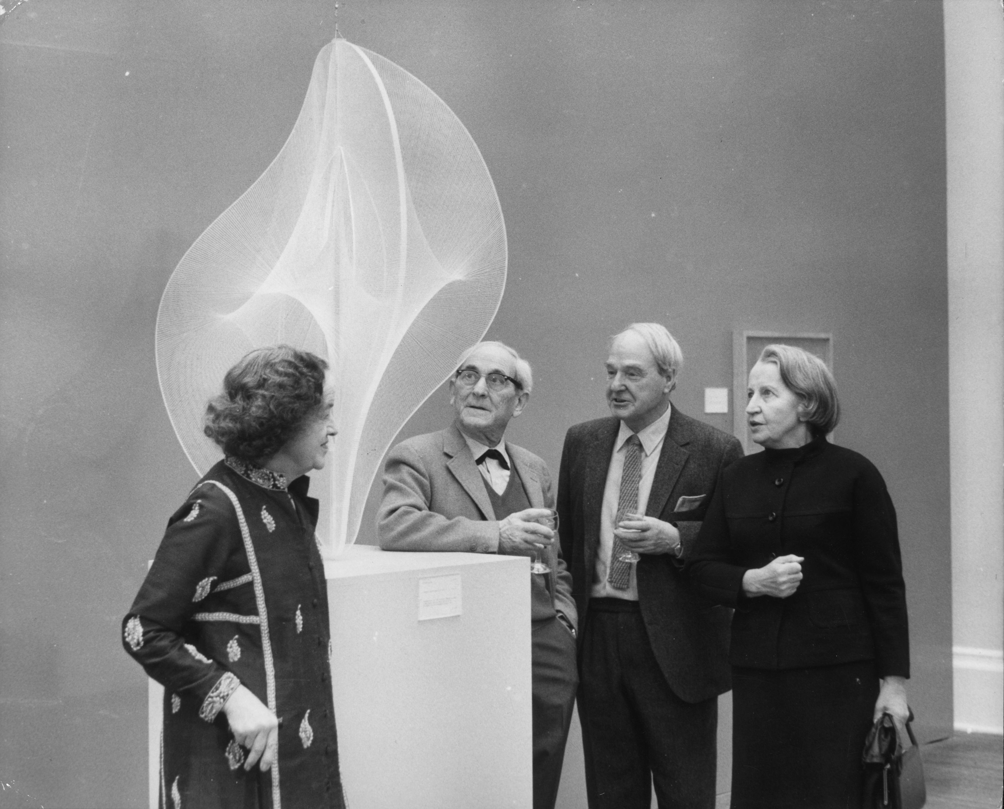 Four individuals examine an abstract sculpture displayed on a pedestal in an art gallery.